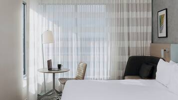 Habitación de hotel con una cama, dos sillas, una mesita con una computadora portátil y una taza, y una ventana con cortinas translúcidas.