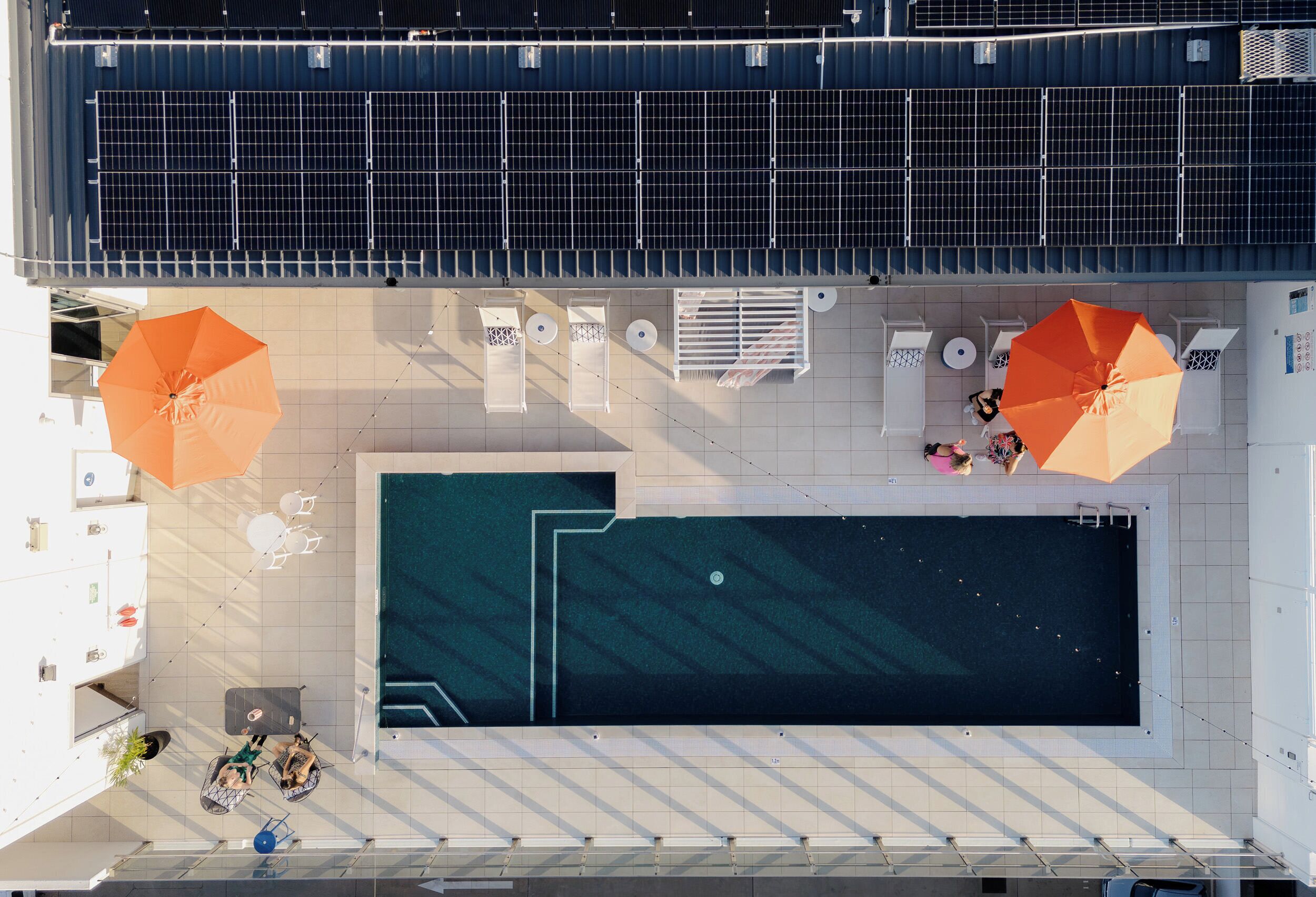 Vista aérea de una zona de piscina en la azotea con paneles solares, dos sombrillas grandes y áreas de estar.