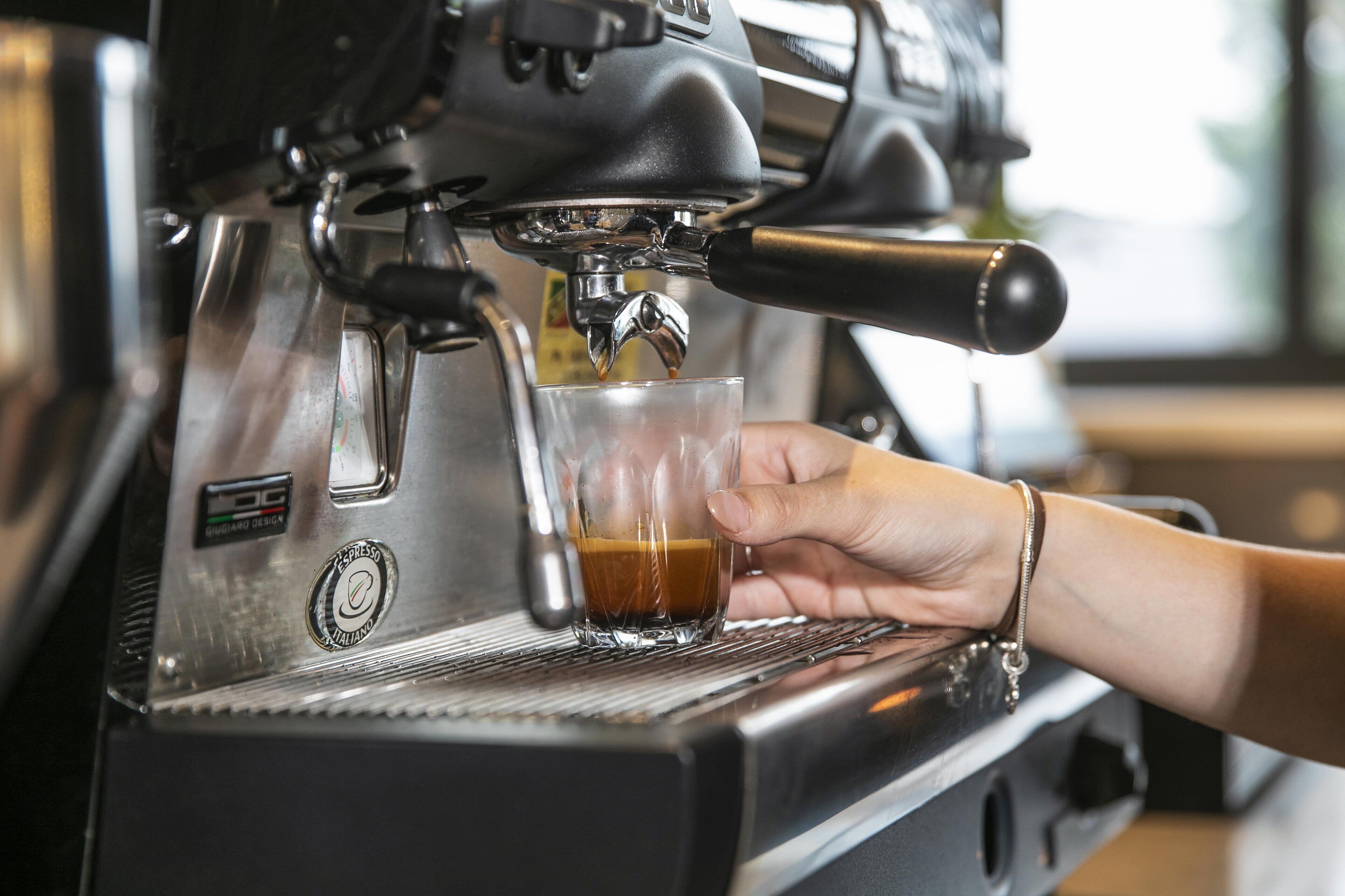 En hånd holder et glas under en espressomaskine, med et logo der viser at det er en maskine fra et bestemt mærke.
