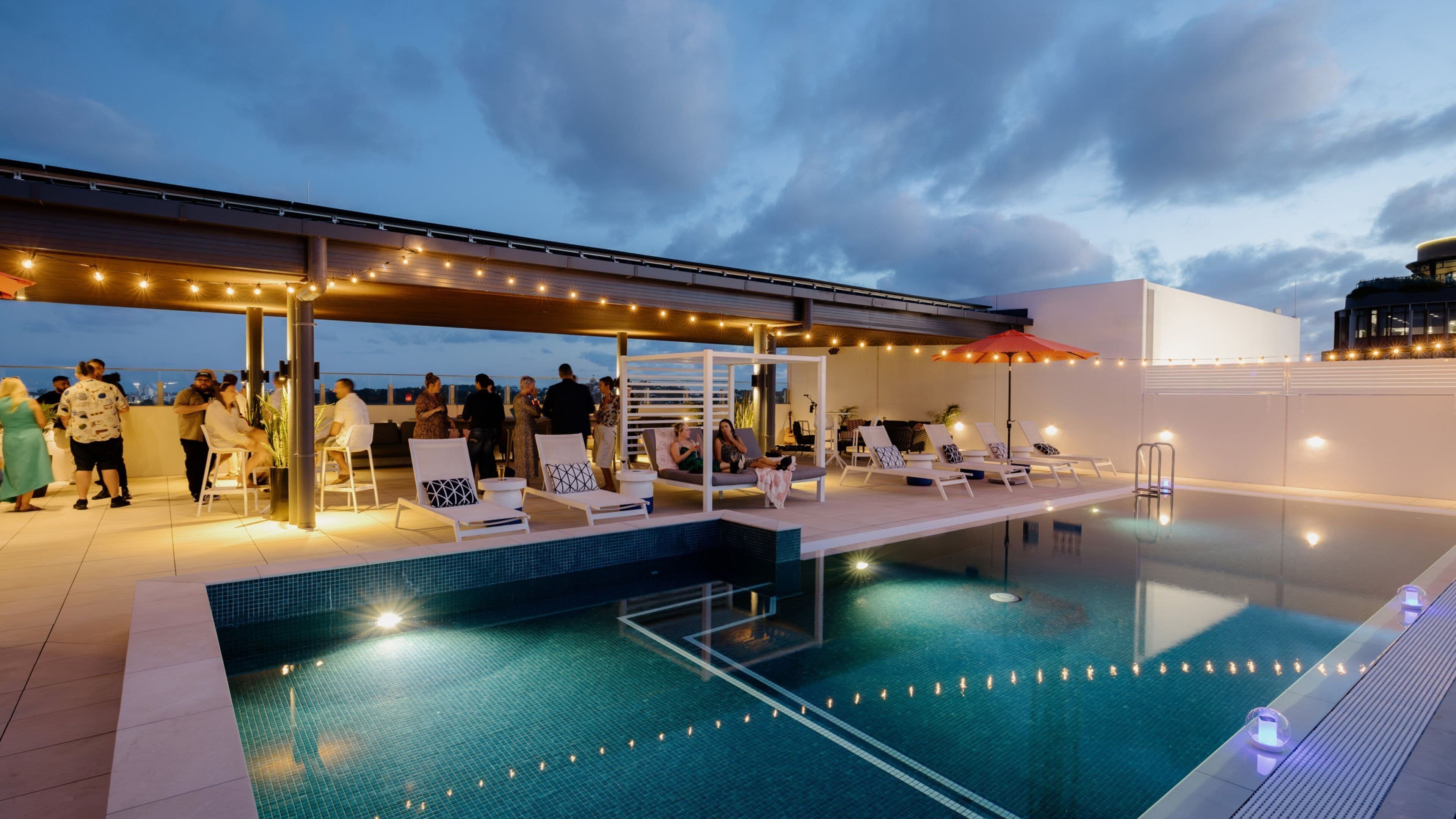 A rooftop pool area with lounge chairs and a covered seating area, surrounded by string lights.