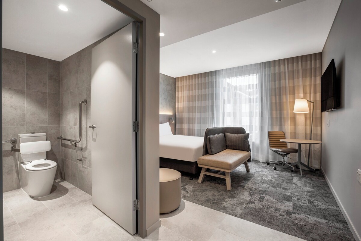 A modern hotel room with a shower, toilet, bed, seating area, and a flat-screen TV mounted on the wall.