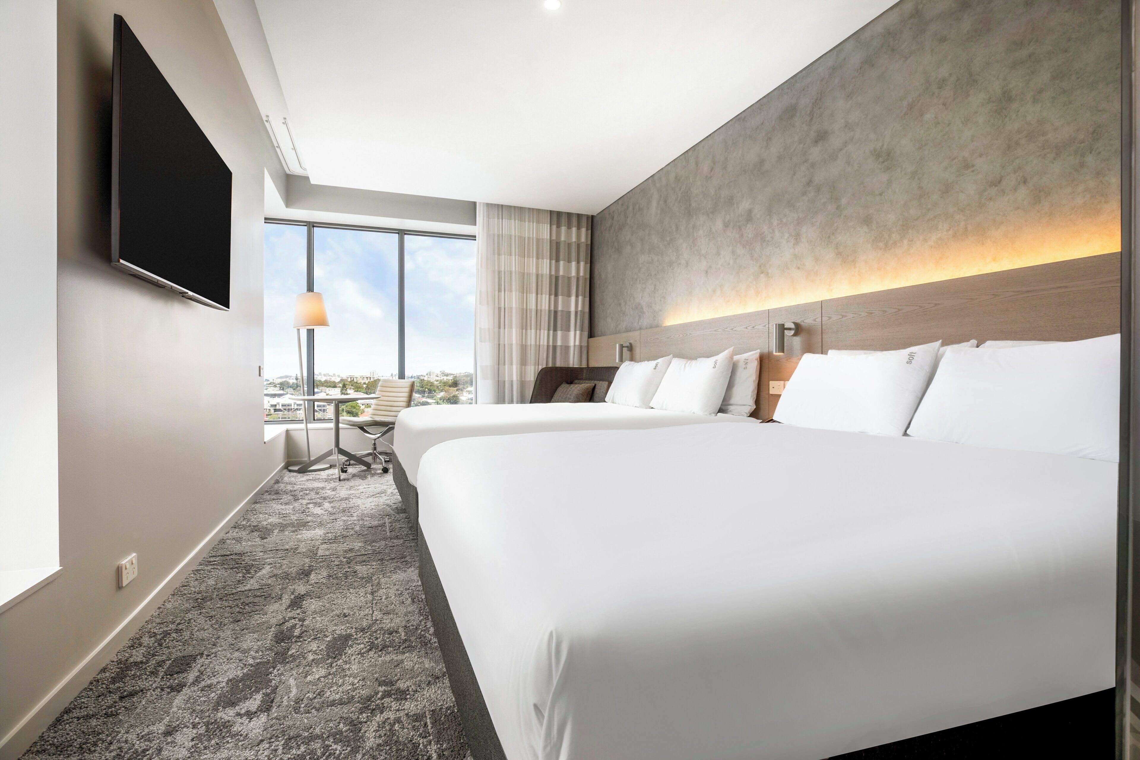 A hotel room with a large bed, a desk, and a chair. There is a TV mounted on the wall and a window with a view of the city.