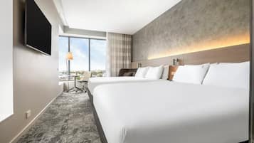 A hotel room with a large bed, a desk, and a chair. There is a TV mounted on the wall and a window with a view of the city.