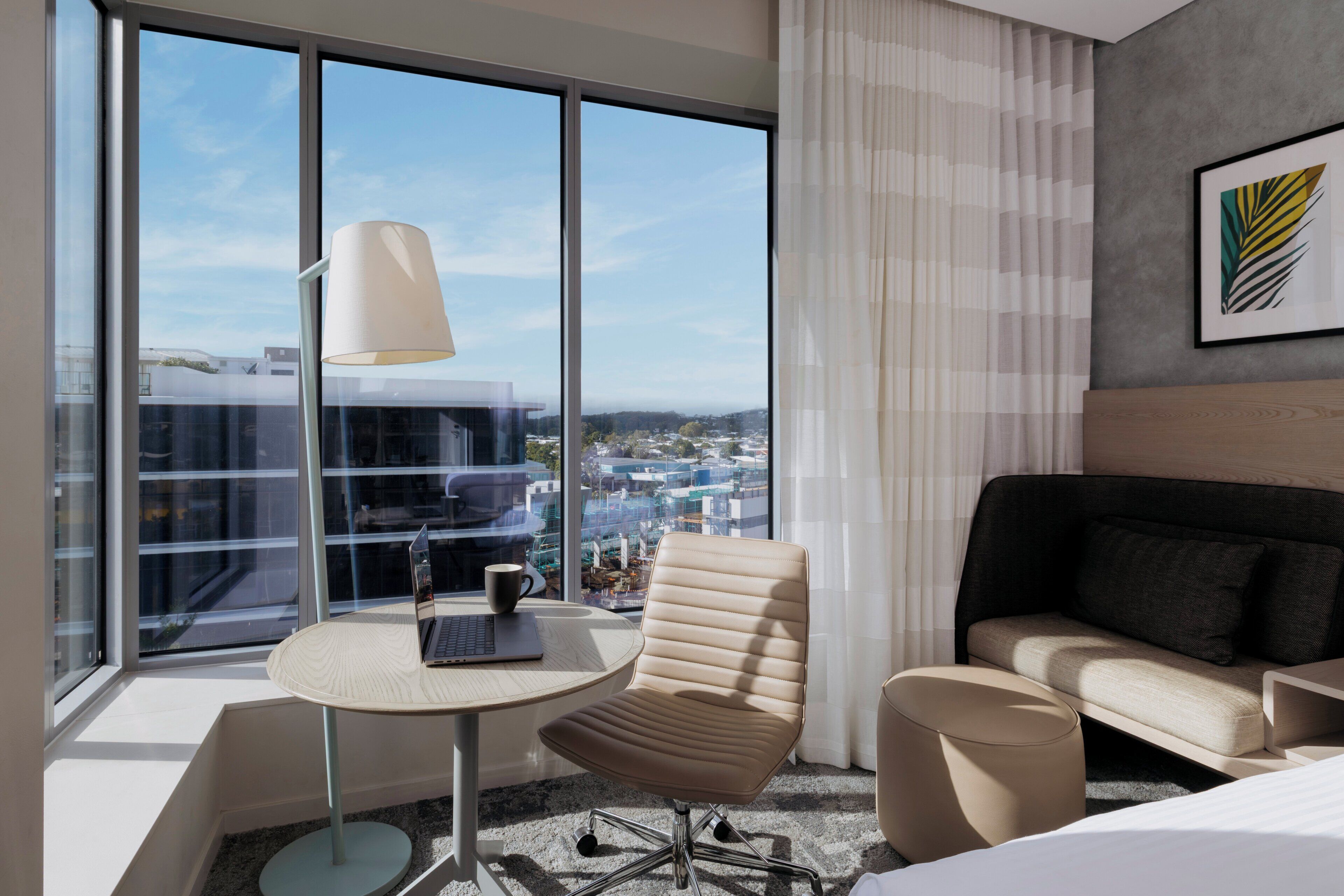 A modern hotel room with a large window, a round table, a cushioned chair, a sofa, and a lamp.