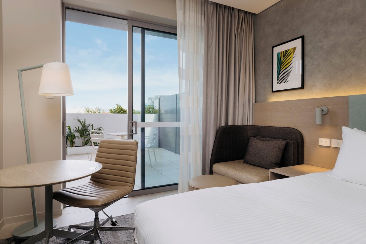 Una habitación de hotel con una cama, una silla, una mesa, una lámpara, vistas al exterior y un cuadro en la pared.