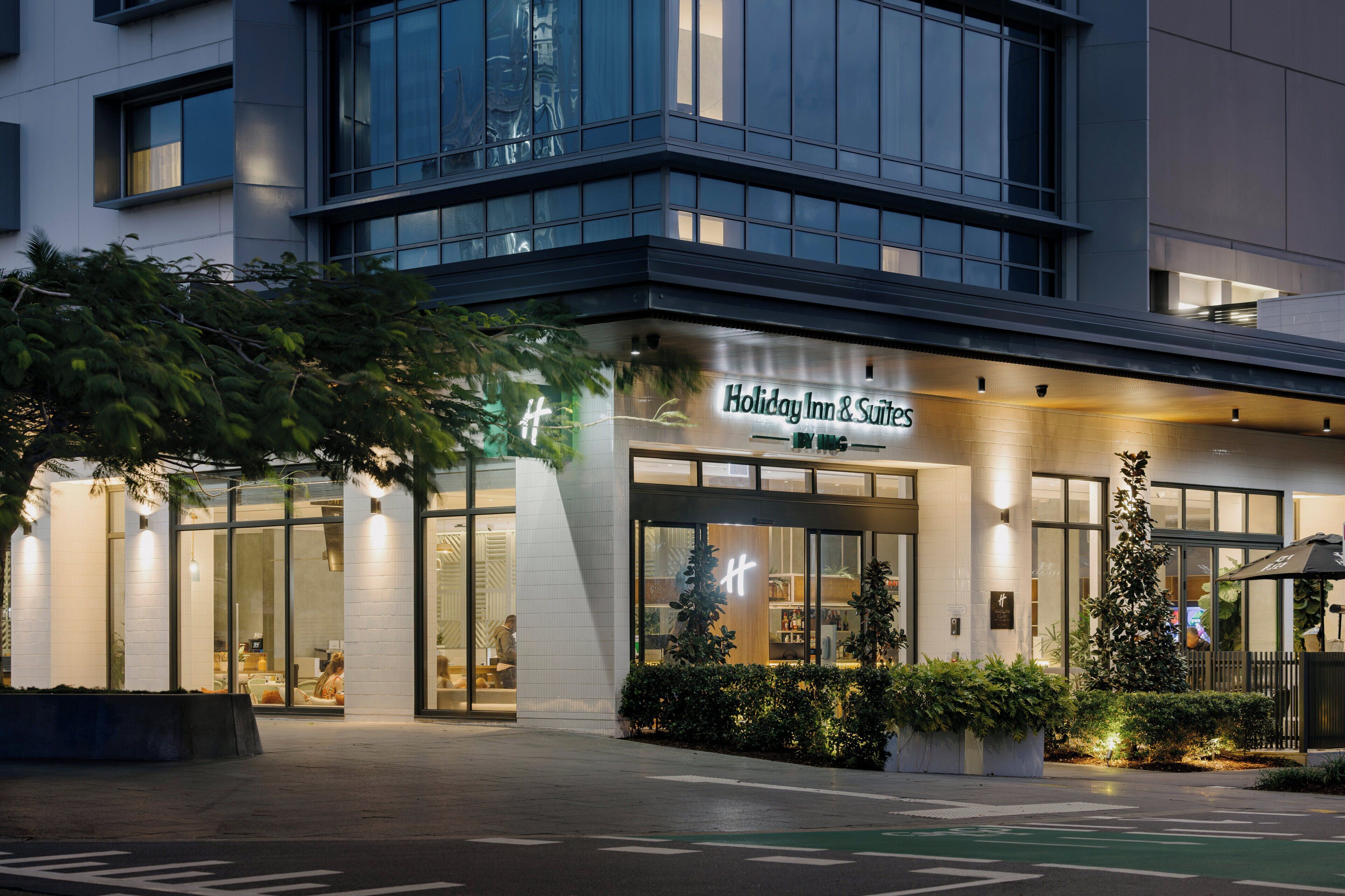 A modern building with a Holiday Inn & Suites sign, large glass windows, and a well-lit entrance.