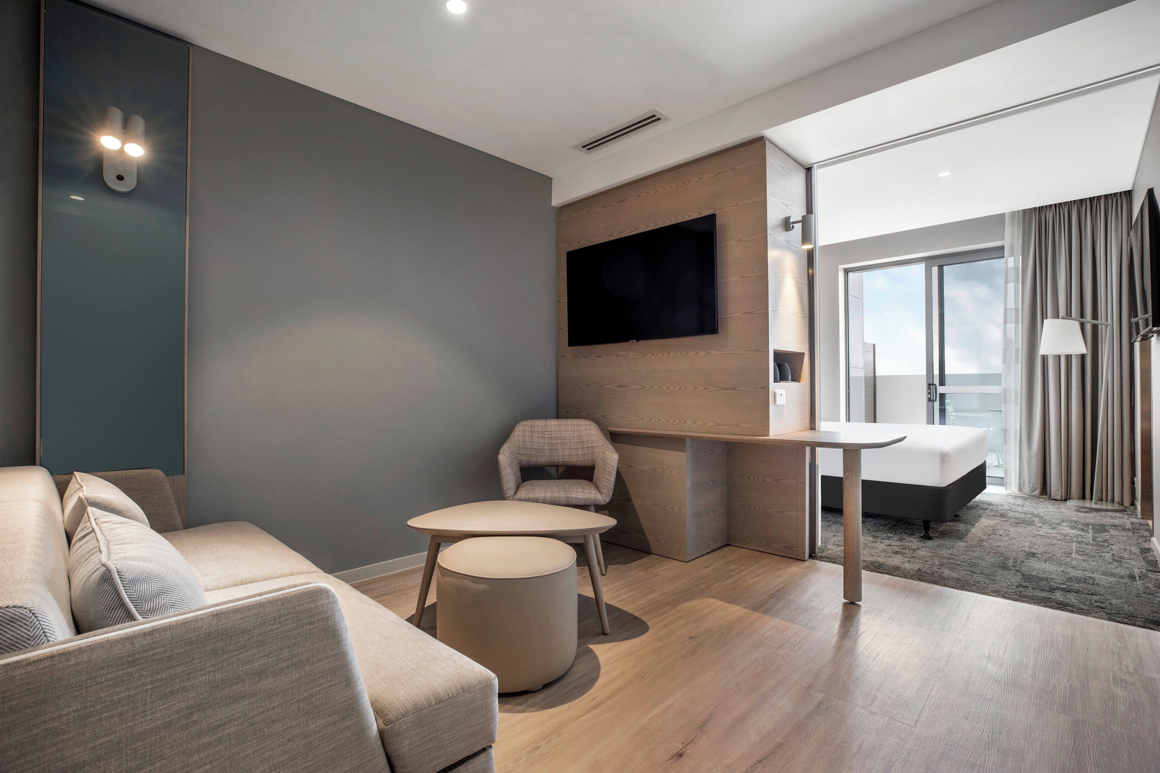 A modern hotel room with a sofa, a small round table, a chair, a flat-screen TV mounted on the wall, and a large window with curtains.