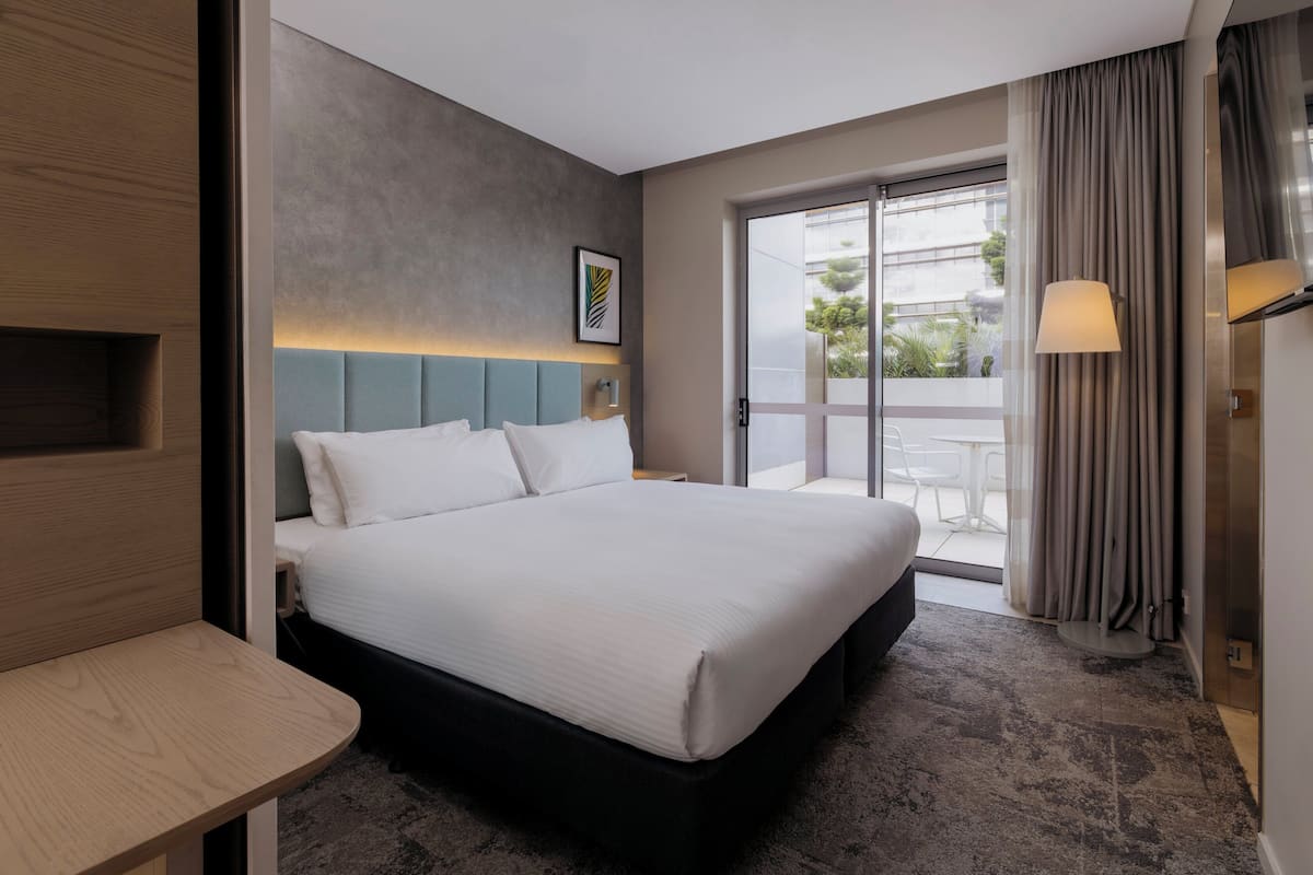 Hotellihuone, jossa on suuri sänky, yöpöytä, seinään kiinnitetty valaisin ja näkymä ulos liukuoven kautta.