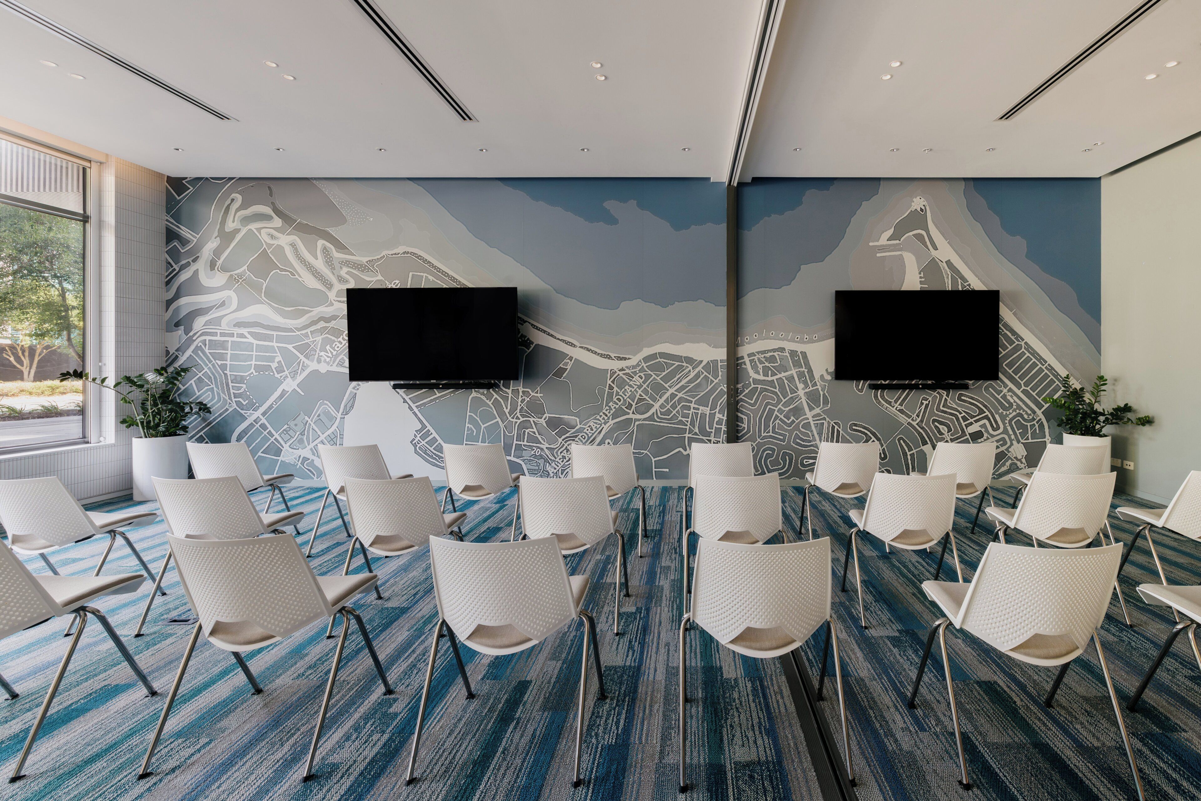 Uma sala de conferências com um grande mural na parede, cadeiras brancas e duas TVs de tela plana.