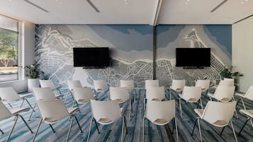 Uma sala de conferências com um grande mural na parede, cadeiras brancas e duas TVs de tela plana.