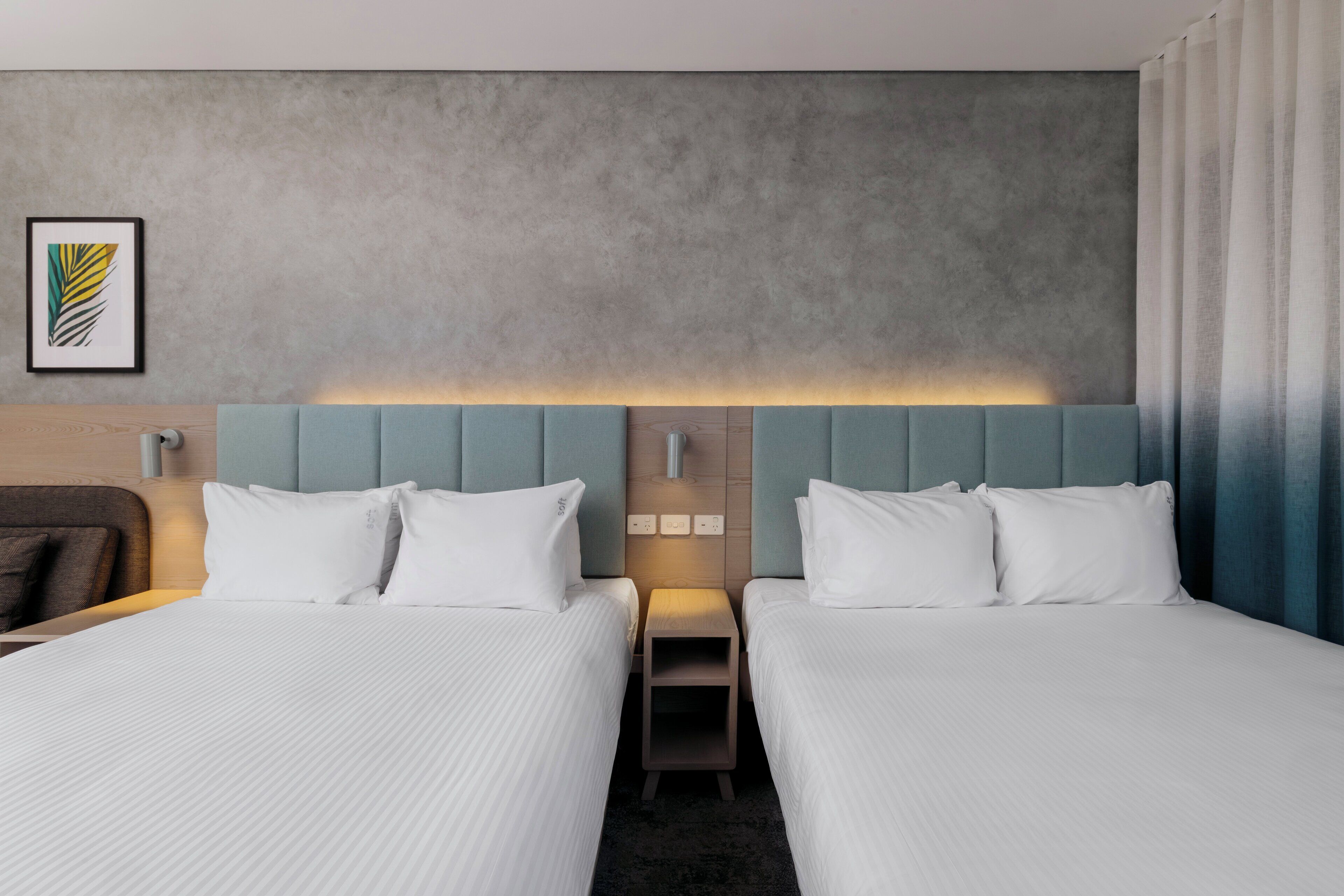 Una habitación de hotel con dos camas, cabecero con panel de luz, mesita de noche pequeña y un cuadro en la pared.