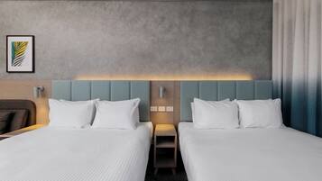 Una habitación de hotel con dos camas, cabecero con panel de luz, mesita de noche pequeña y un cuadro en la pared.