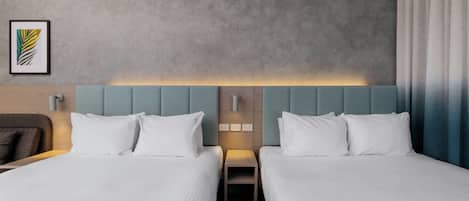 A hotel room with two beds, a headboard with a light panel, a small bedside table, and a framed picture on the wall.