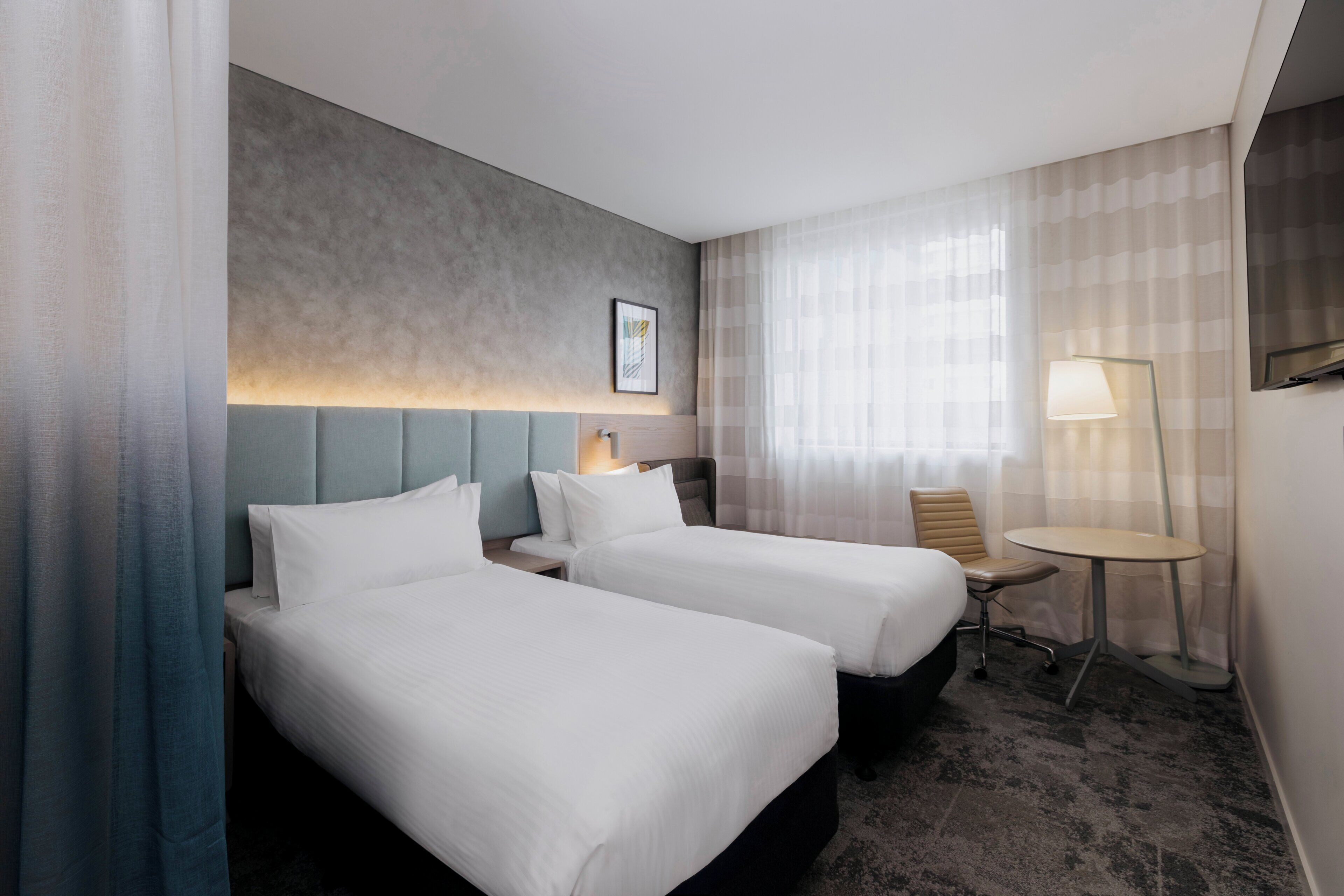 A hotel room with two beds, a desk, a chair, a lamp, and a window with curtains.