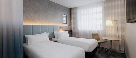 A hotel room with two beds, a desk, a chair, a lamp, and a window with curtains.