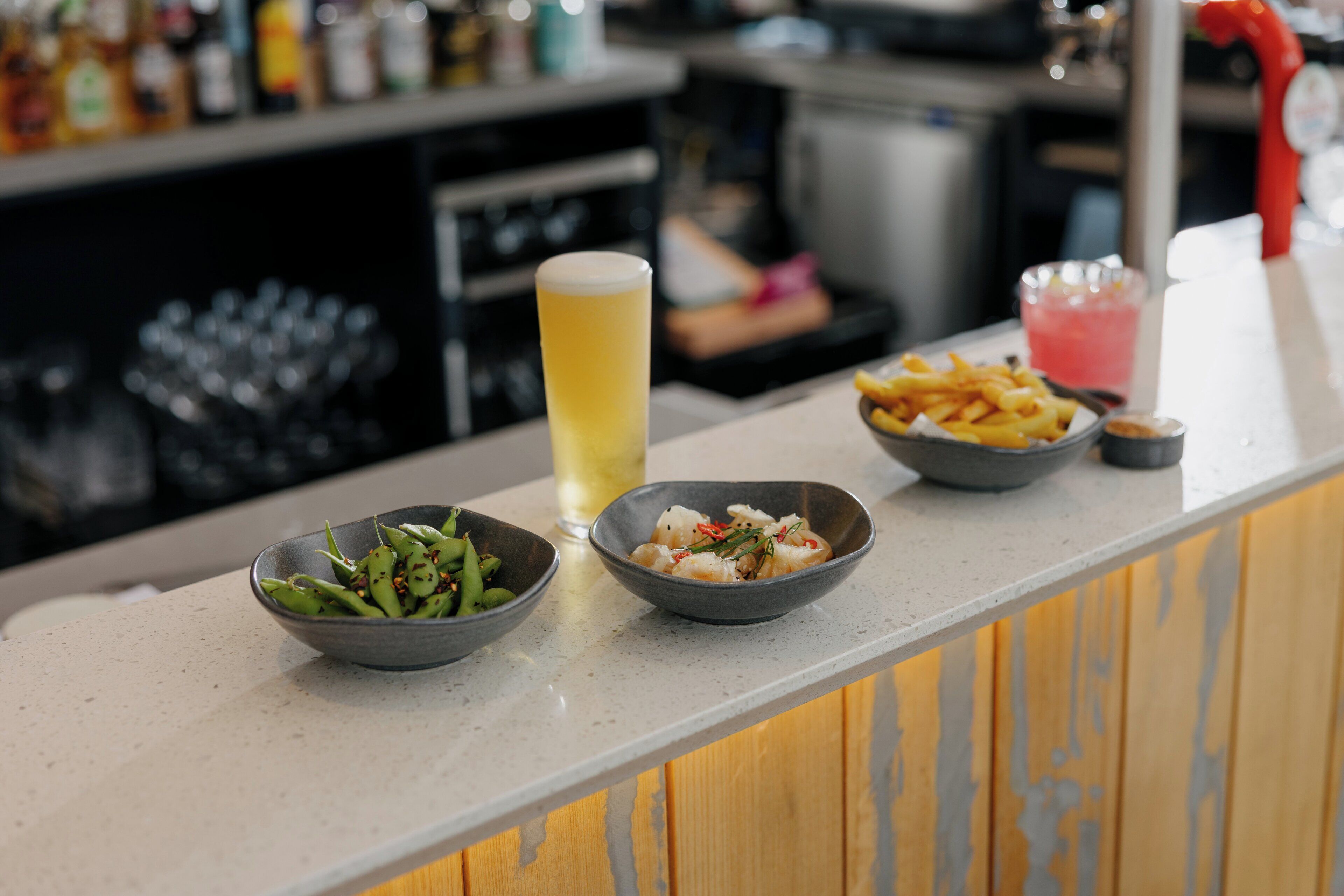 A bar counter with a tall glass of beer, two bowls of food, and a pink drink.