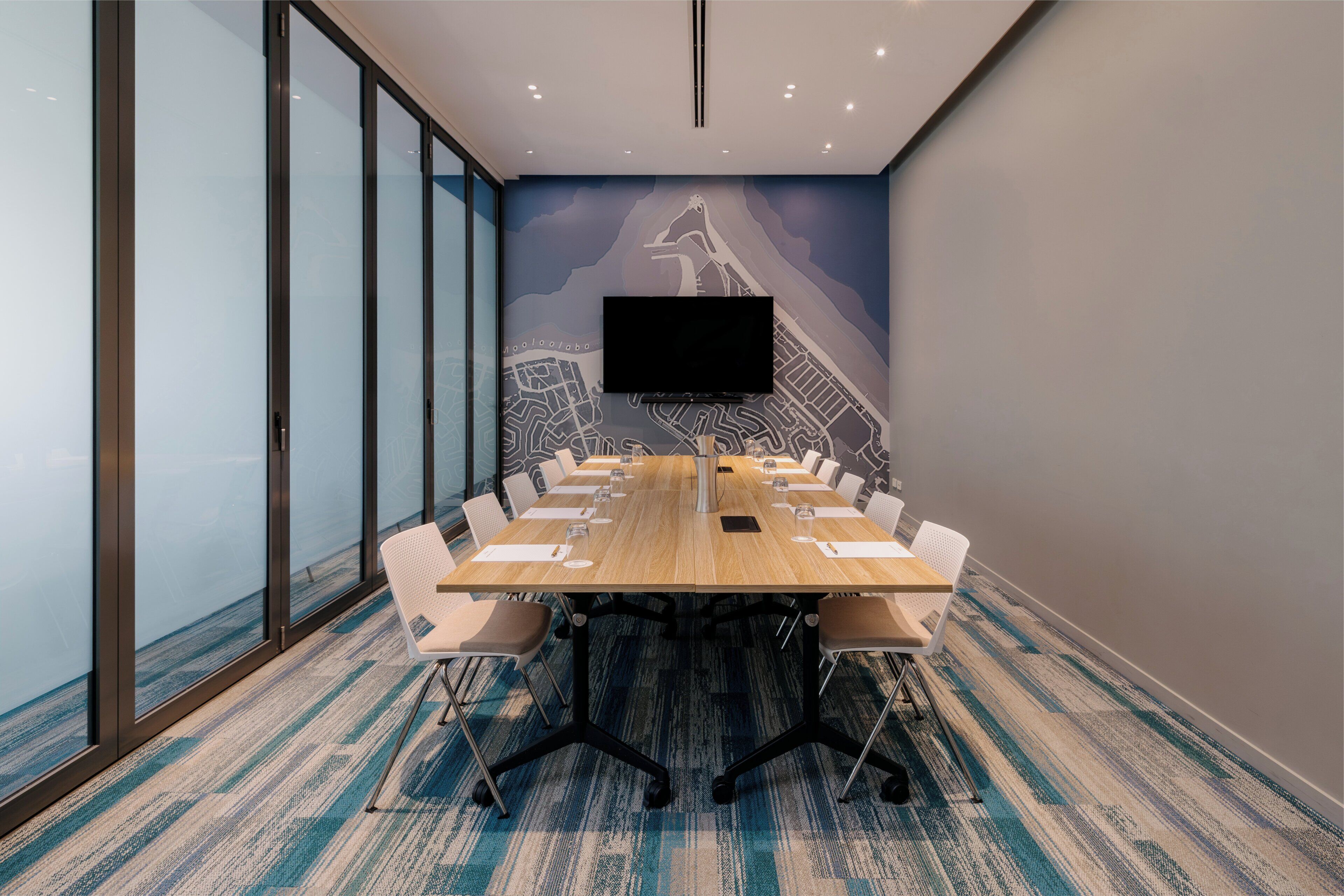 A conference room with a long table, white chairs, a flat-screen TV, and a cityscape mural on the wall.