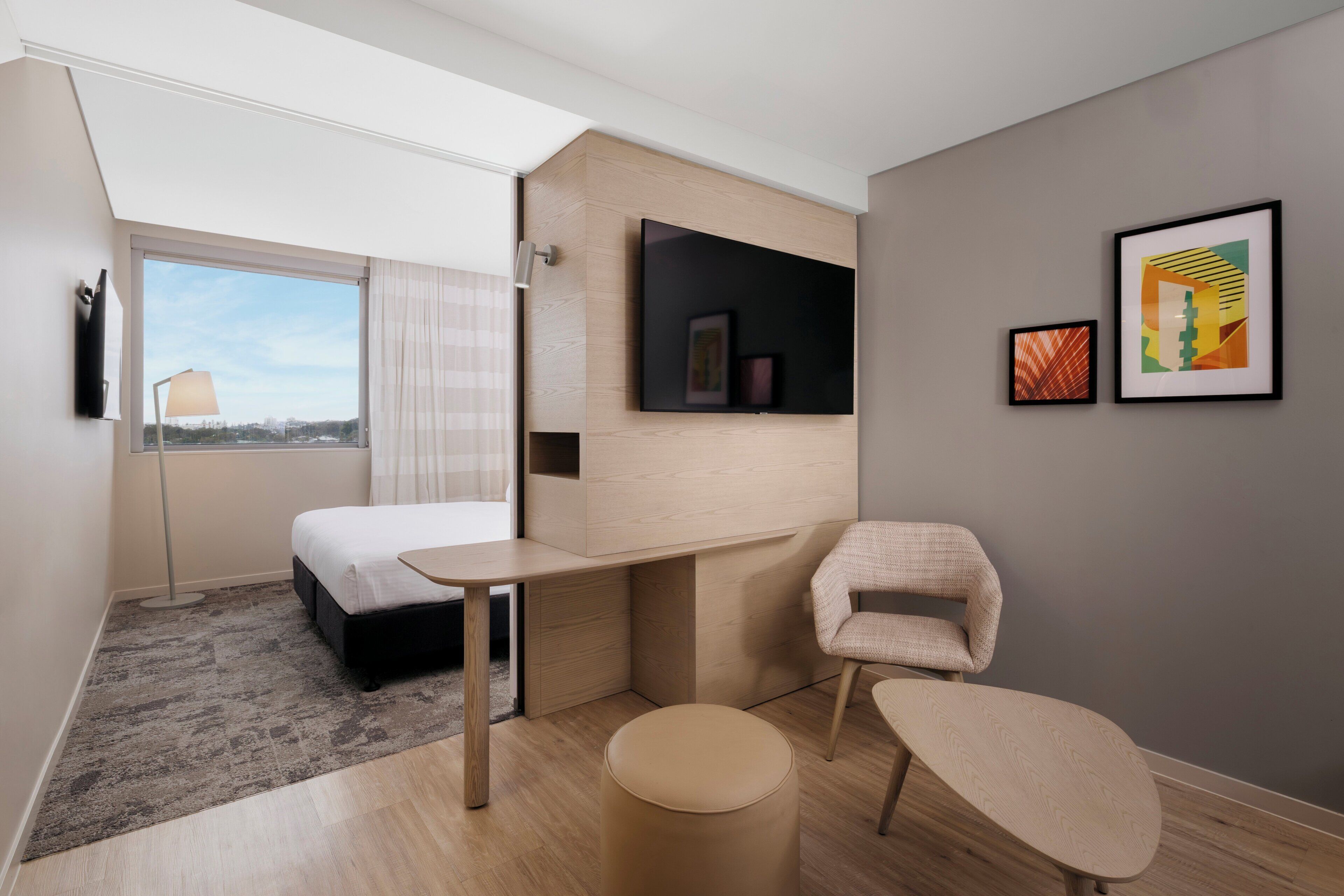 A modern hotel room with a bed, desk, chair, and artwork on the wall.