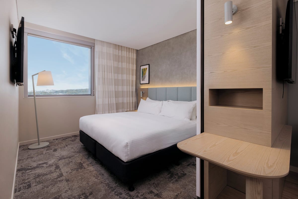 Hotellihuone, jossa on sänky, yöpöytä, televisio, ikkuna verhoilla ja seinälle kiinnitetty hylly.