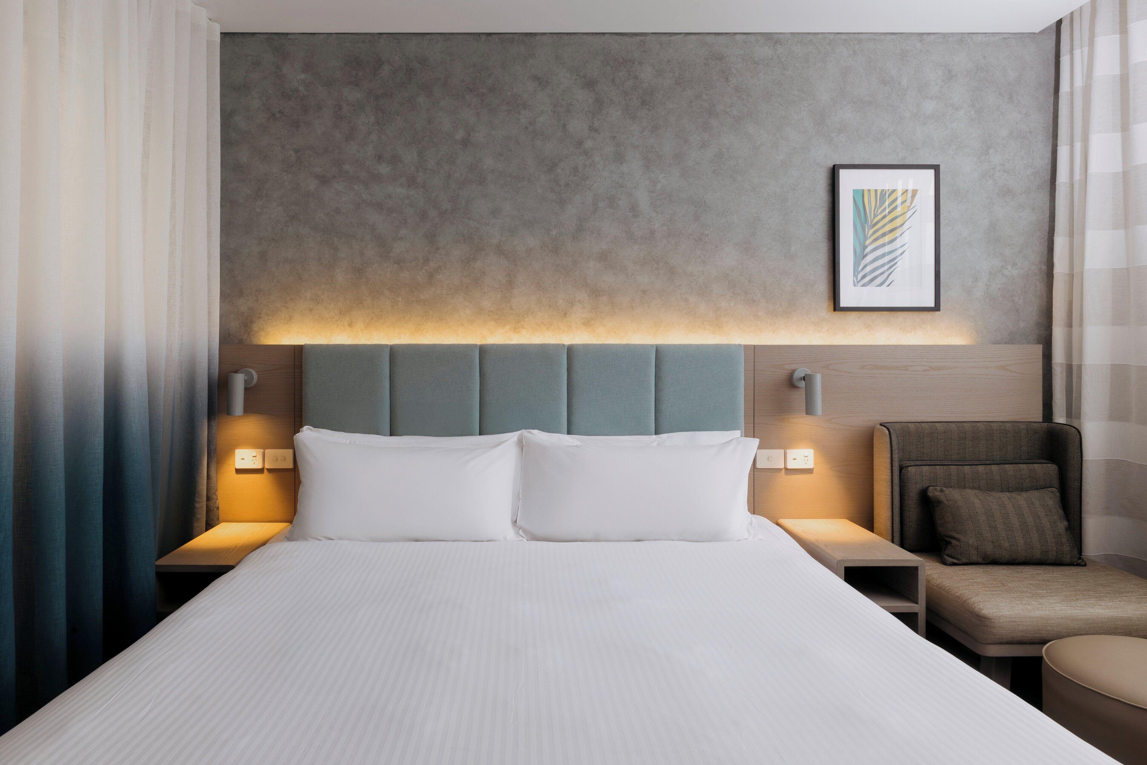 Una habitación de hotel con una cama grande, dos mesitas de noche con lámparas, un cabecero, un cuadro enmarcado en la pared y una zona de estar con un sillón y reposapiés.