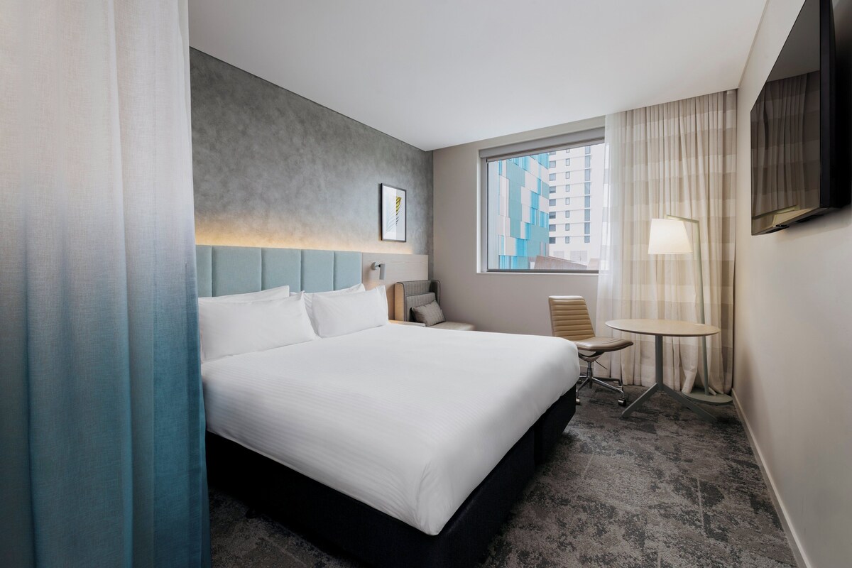 Una habitación de hotel con una cama grande, un escritorio, una silla, una lámpara, un televisor y una ventana con vistas a la ciudad.