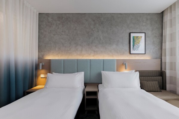 Habitación de hotel con dos camas, cabecero gris, una obra de arte enmarcada y una lámpara de noche.
