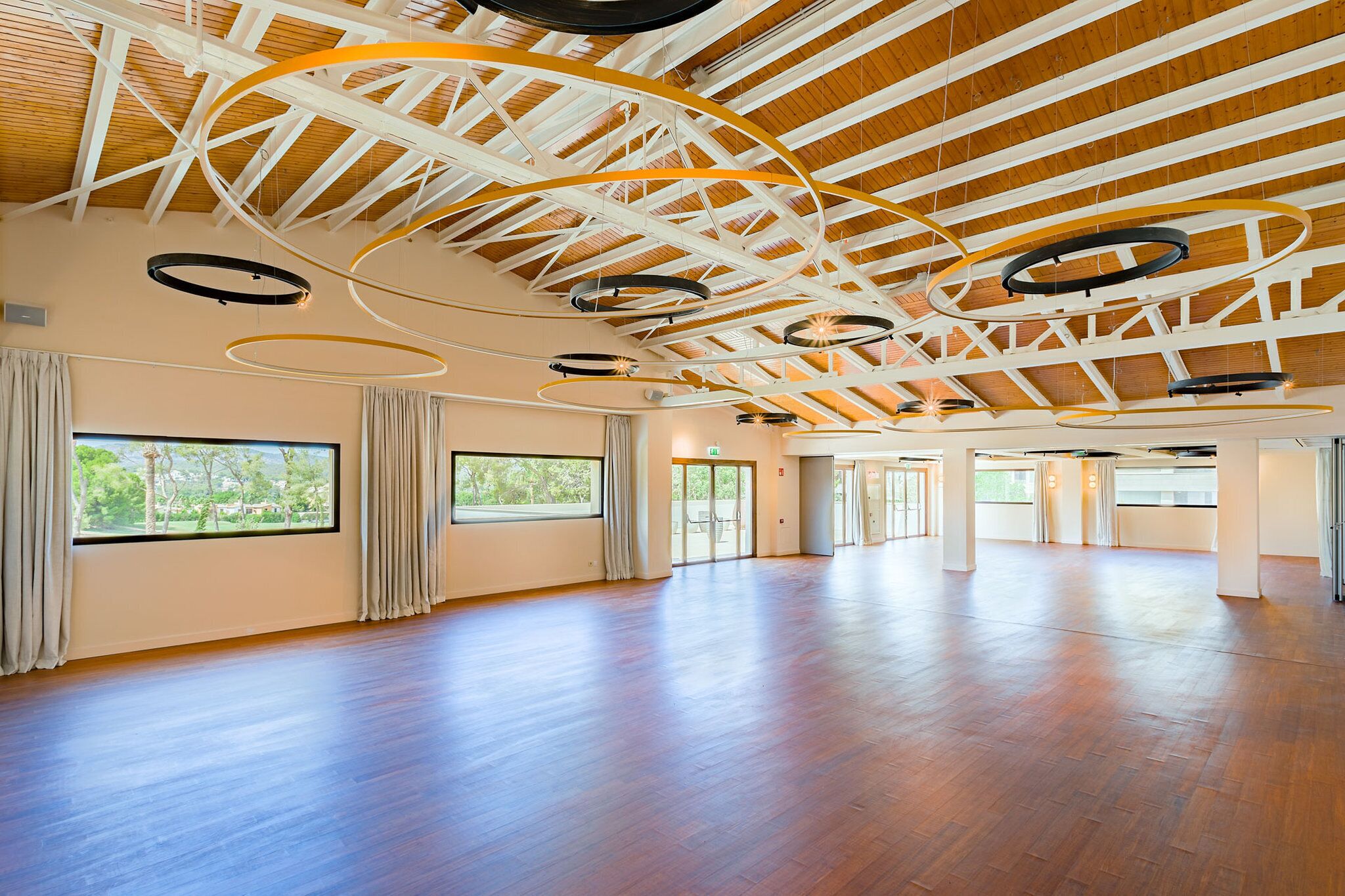 A spacious room with wooden floors, large windows, and a high ceiling with exposed beams and circular light fixtures.
