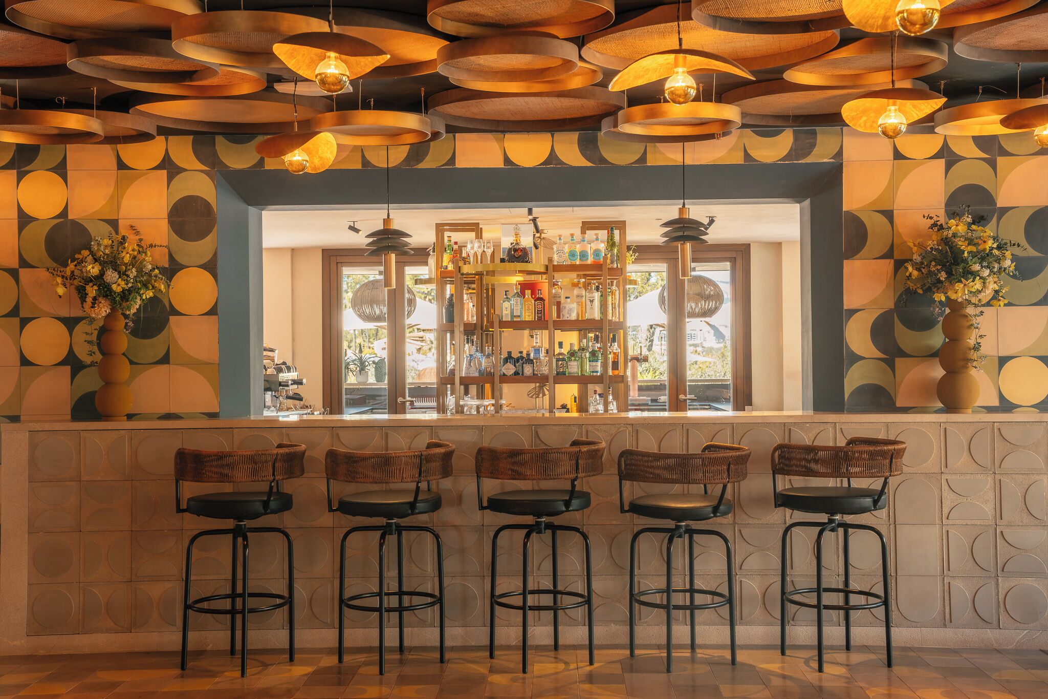 Un espace bar avec un mur décoré de motifs, une étagère remplie de bouteilles et des tabourets de bar en osier.
