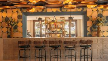 Un espace bar avec un mur décoré de motifs, une étagère remplie de bouteilles et des tabourets de bar en osier.