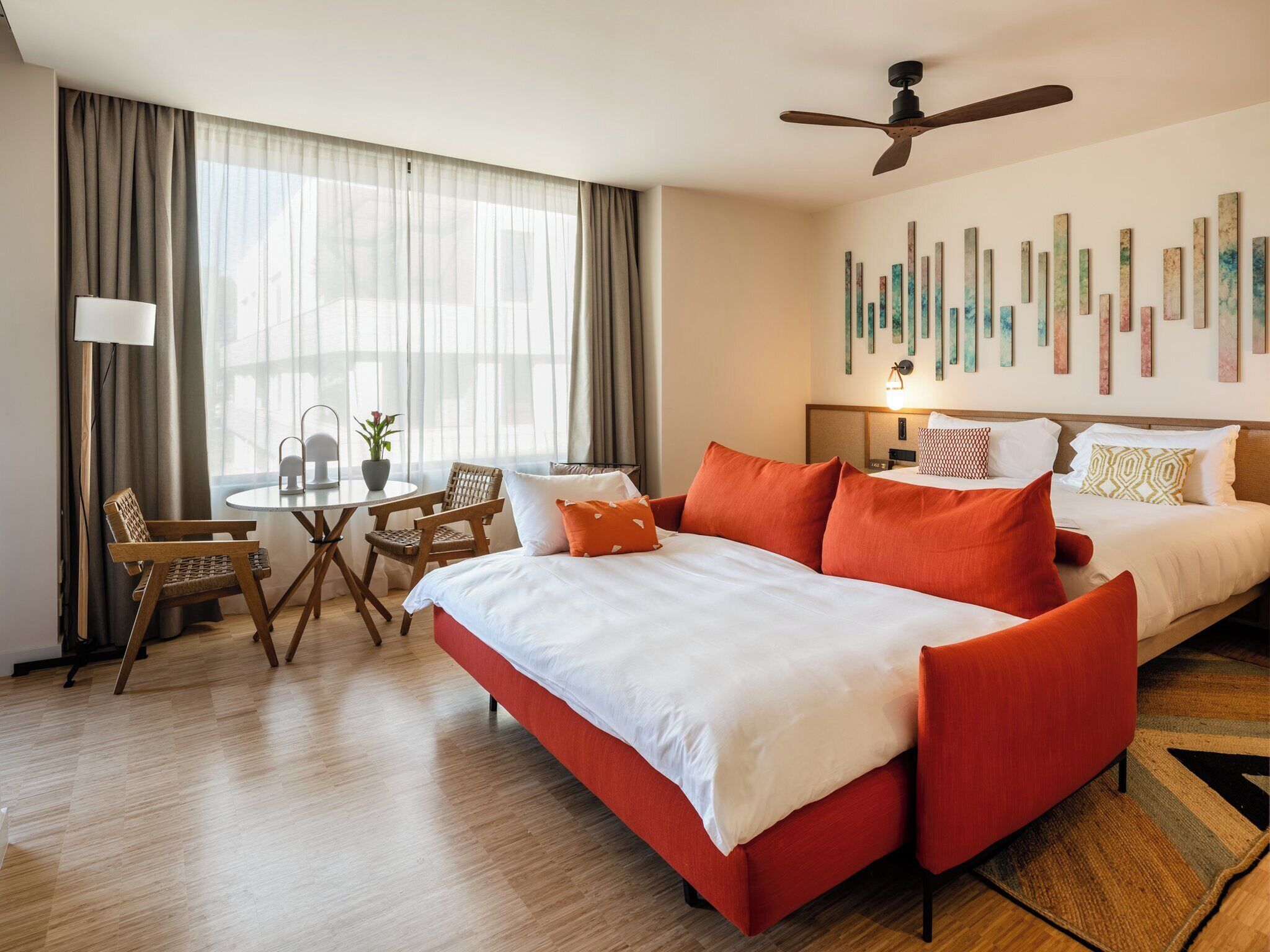 A hotel room with a bed, a desk, a chair, a ceiling fan, and artwork on the wall.