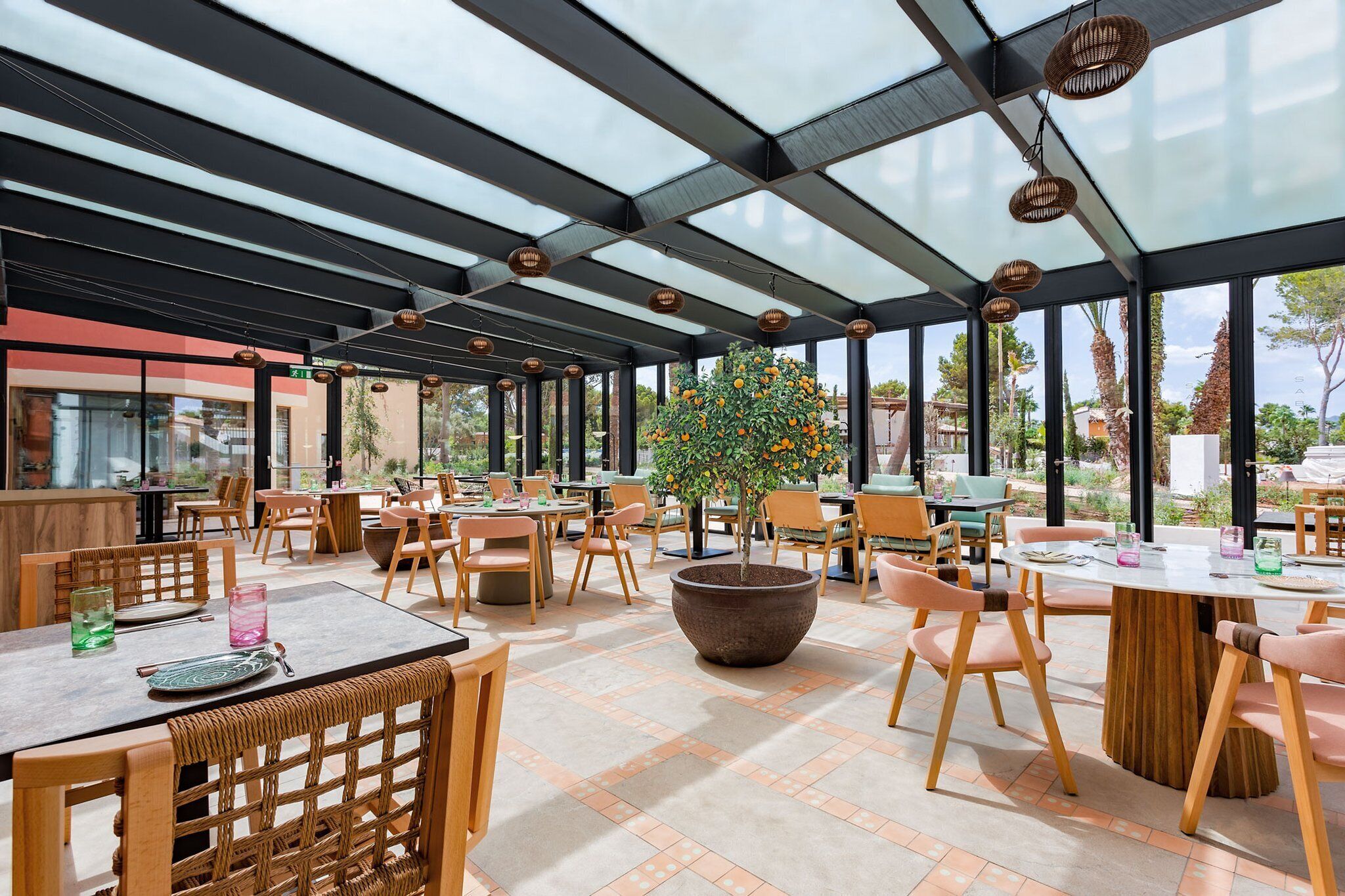 A spacious outdoor seating area with tables and chairs, a large planter with a tree, and a glass ceiling structure.