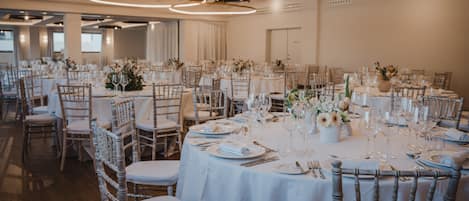 A large banquet hall with round tables set for a formal event, featuring white tablecloths, floral centerpieces, and wooden chairs.