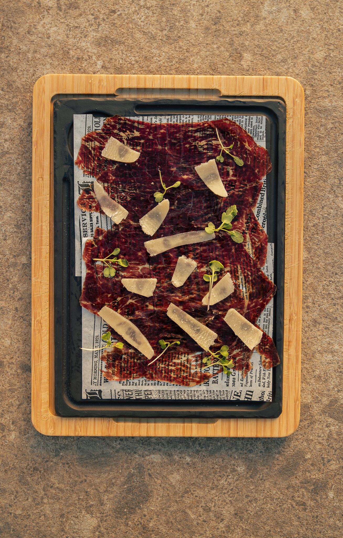 Un plateau en bois recouvert d’un journal, contenant des tranches de charcuterie et de fromage, et agrémenté d’herbes aromatiques.