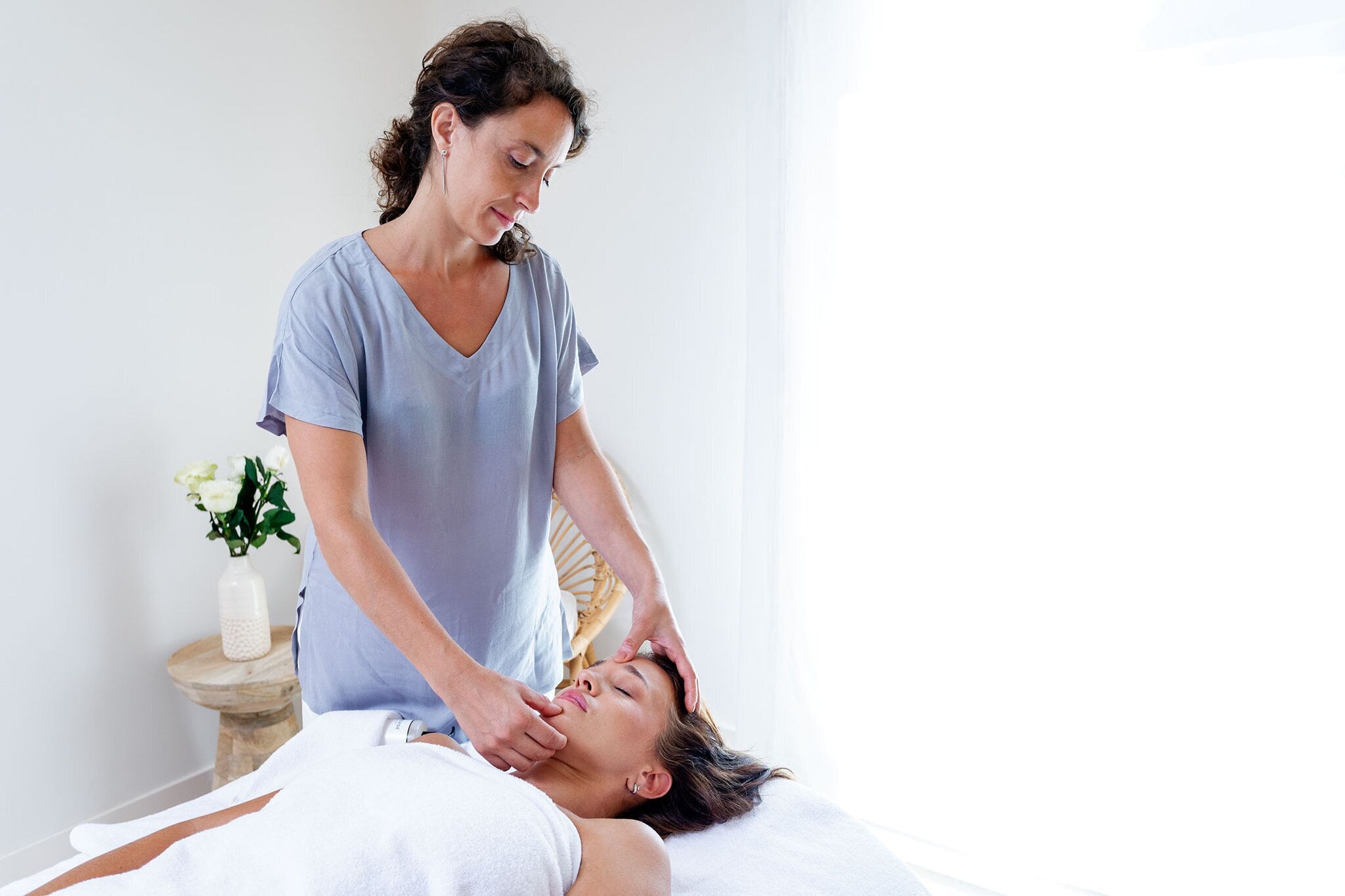 A person receiving a massage in a room with a bed, a small table with a vase of flowers, and a white wall.