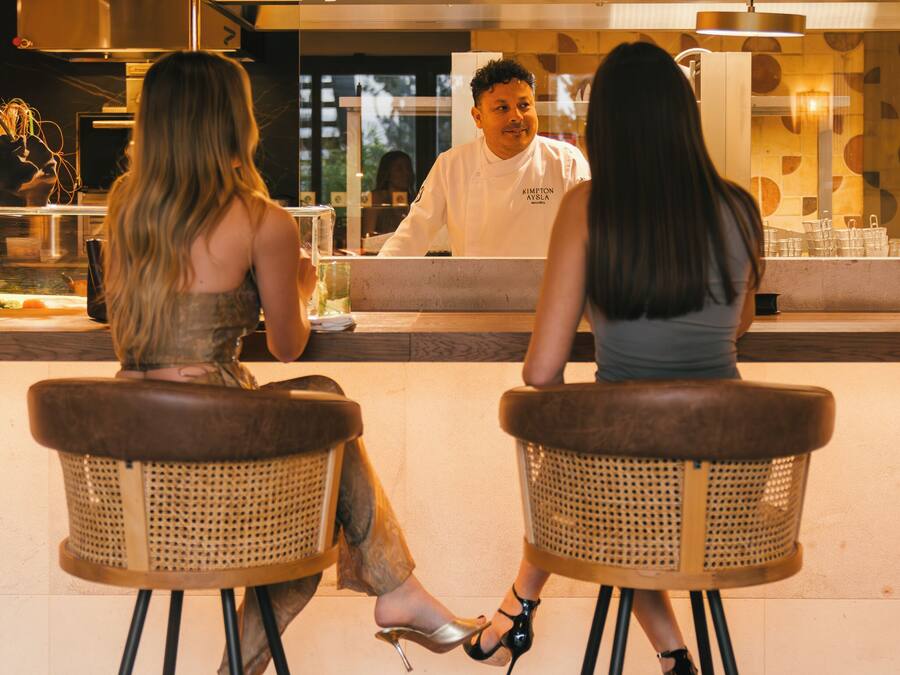 Dos personas sentadas en una barra con taburetes, frente a una barra donde un camarero las atiende.