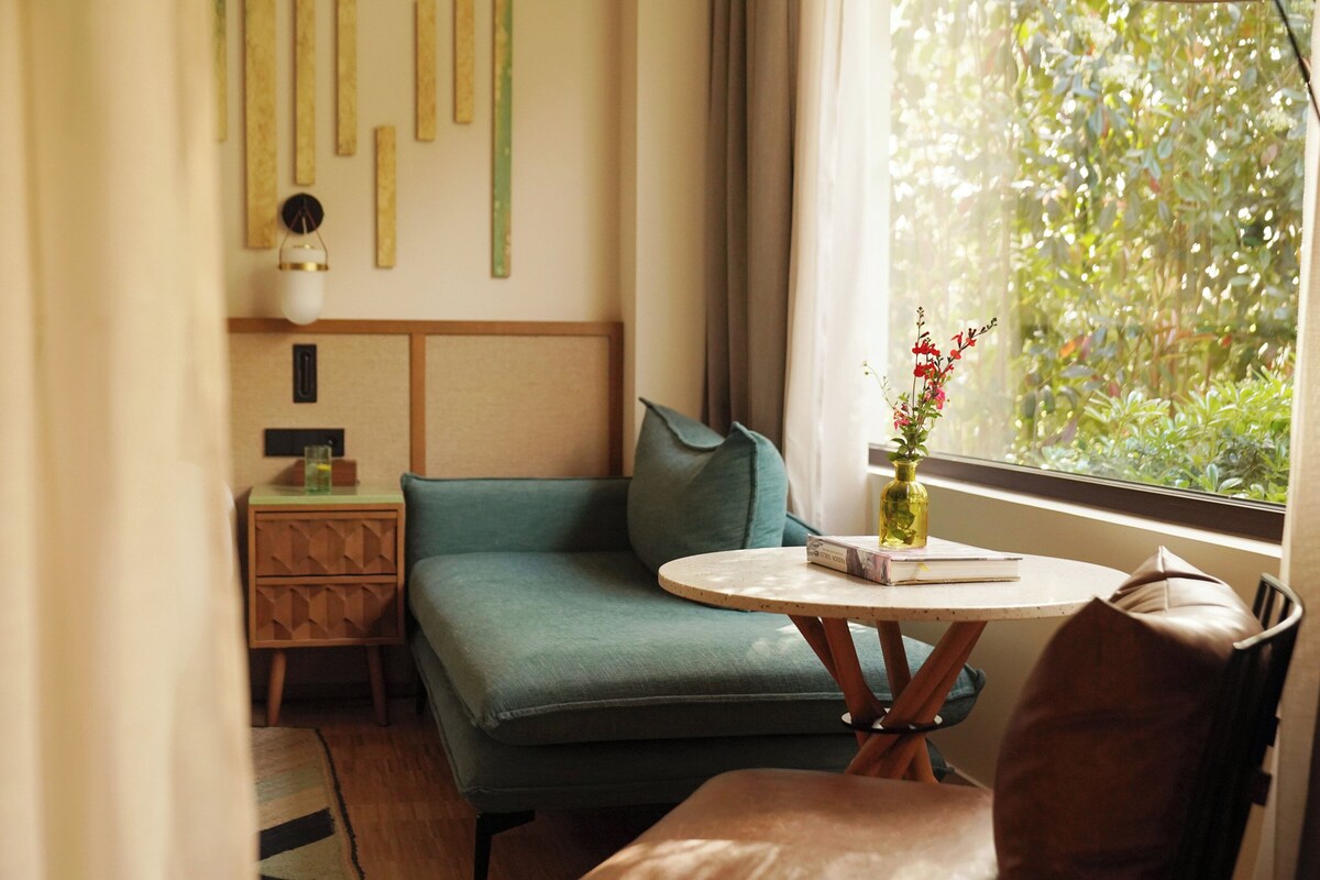 A room with a green sofa, a round table with a vase, a window with a view of greenery, and a wall with vertical wooden elements.