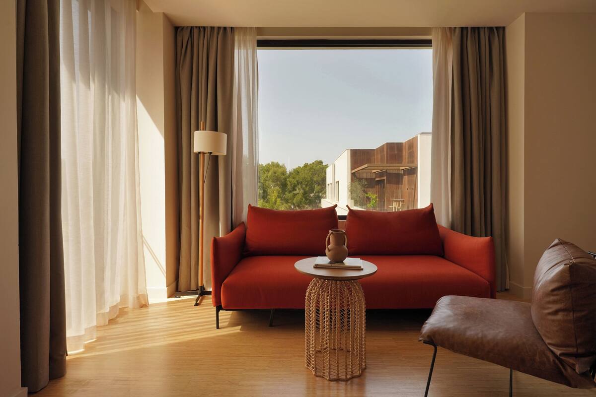 A modern living room with a red sofa, a brown leather chair, a woven side table, and a view of buildings through large windows.
