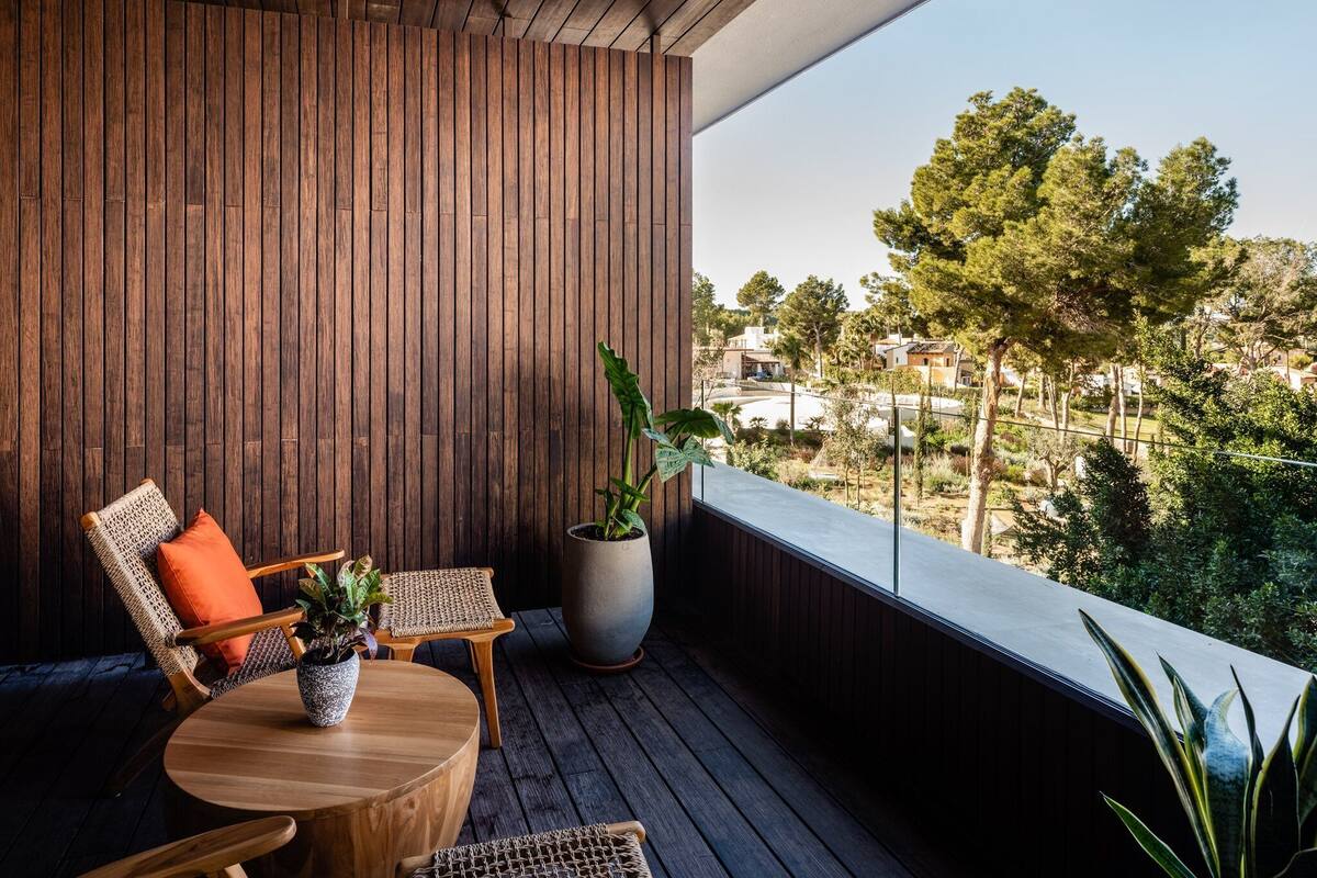 A balcony with a wooden floor, a round wooden table, two wicker chairs, a potted plant, and a view of a residential area with trees and houses.