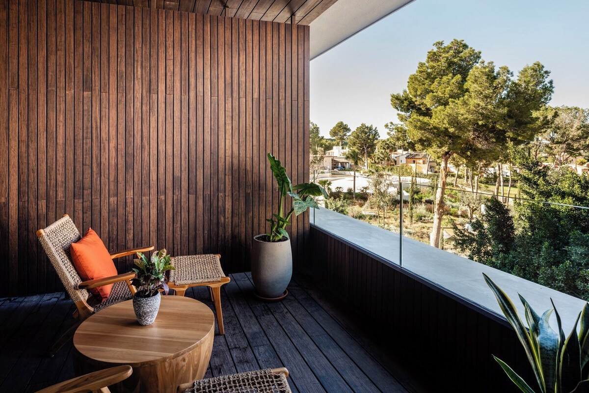 A balcony with wooden flooring, a round table, two wicker chairs, a potted plant, and a view of a residential area with trees and houses.