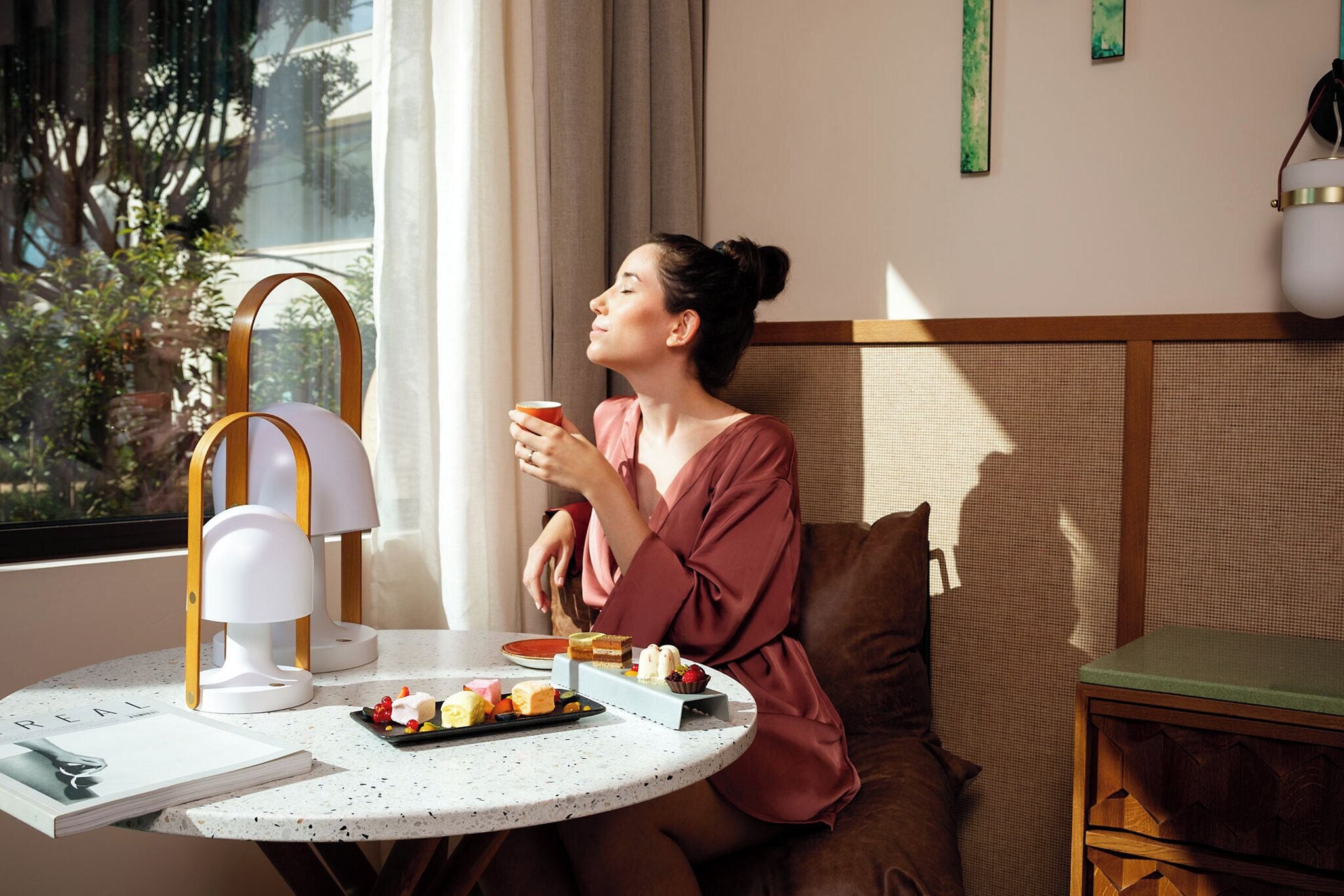 Iemand zit aan een tafel met een lamp, een bord eten en een raam met gordijnen.