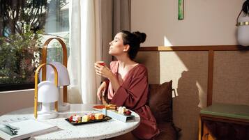 A person seated at a table with a lamp, a plate of food, and a window with curtains.