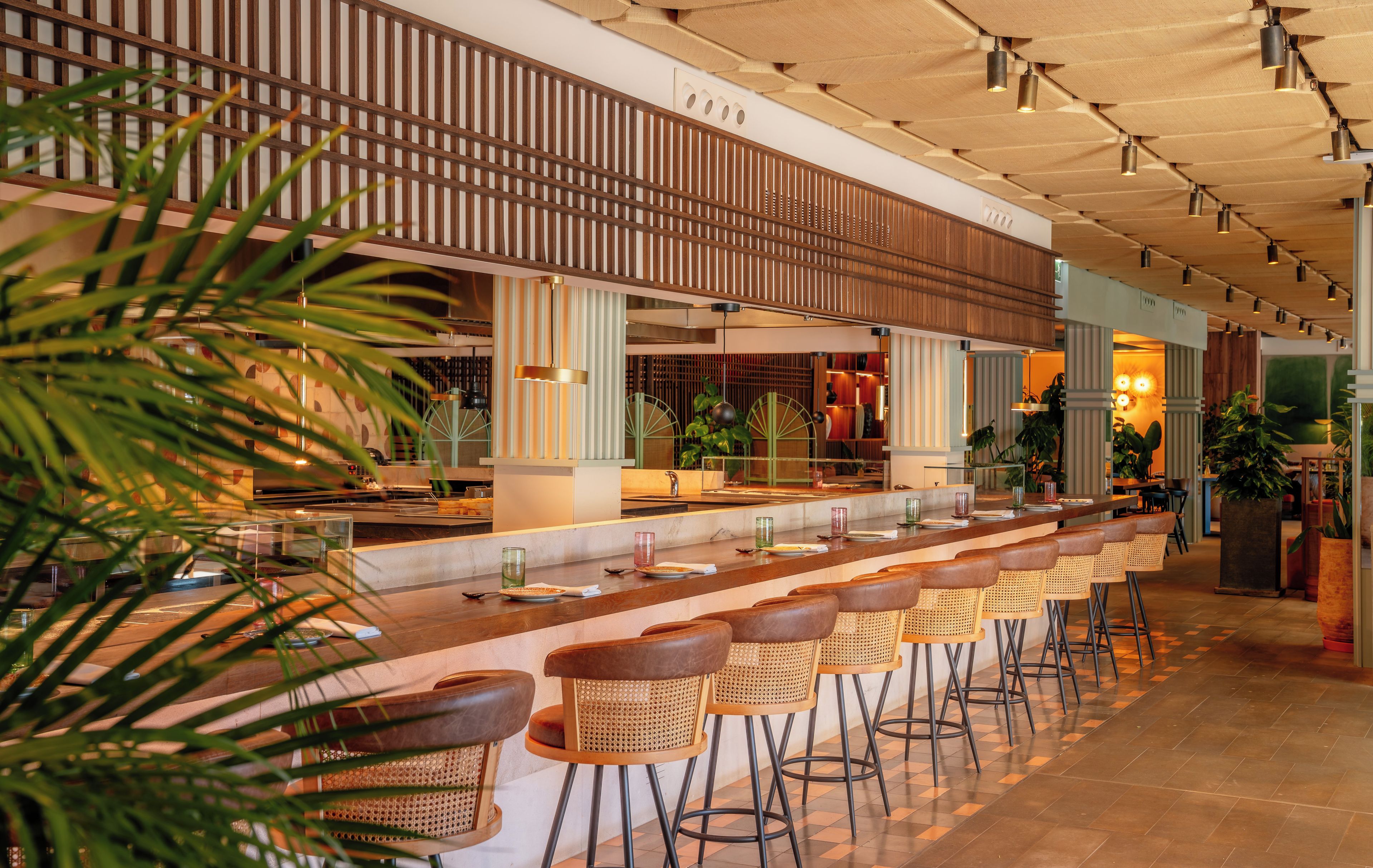 A bar area with a long counter, multiple stools, and a ceiling with wooden slats and hanging lights.