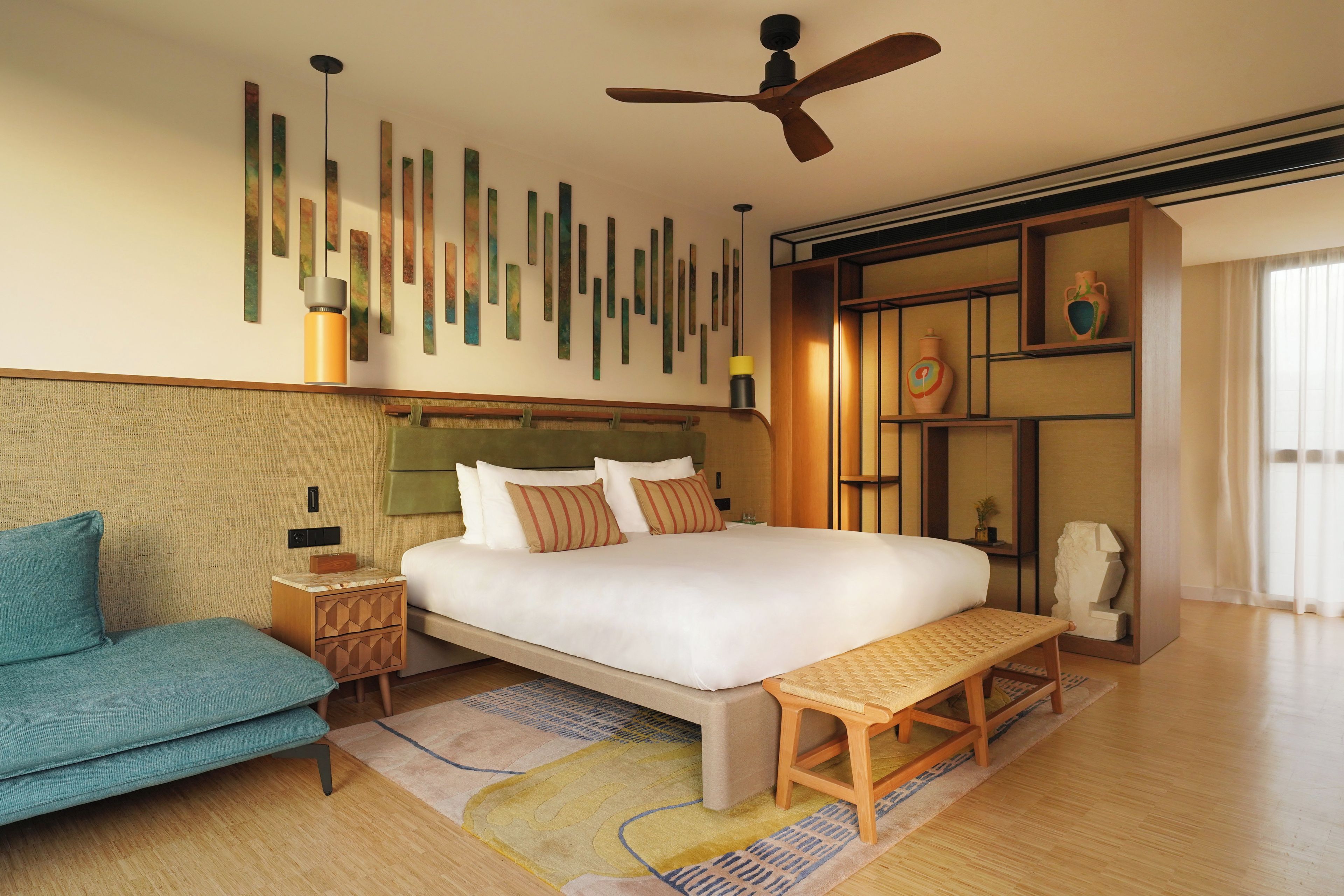 Ein modernes Hotelzimmer mit einem Bett, einer Couch, einer Holzbank, einem Deckenventilator und einer Wand mit abstrakter Kunst.