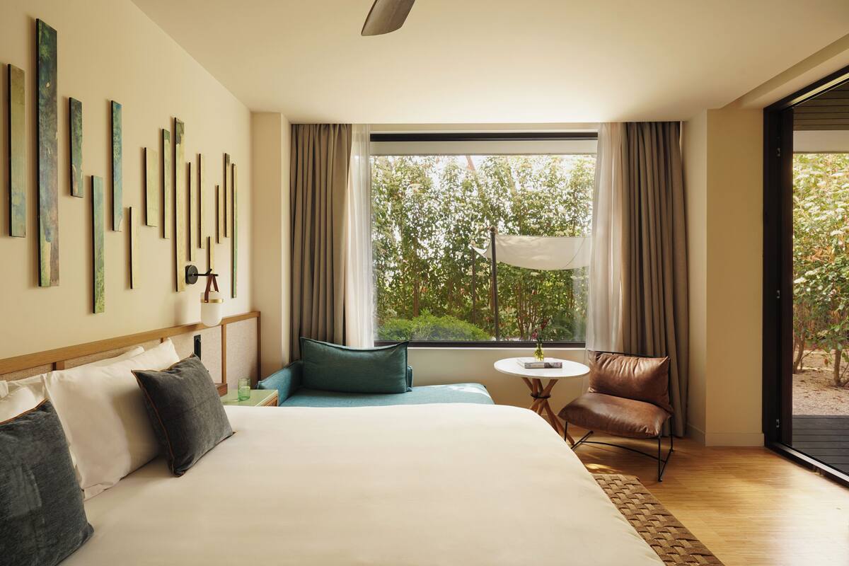 A hotel room with a large bed, a seating area, a small table, and a view of greenery outside.