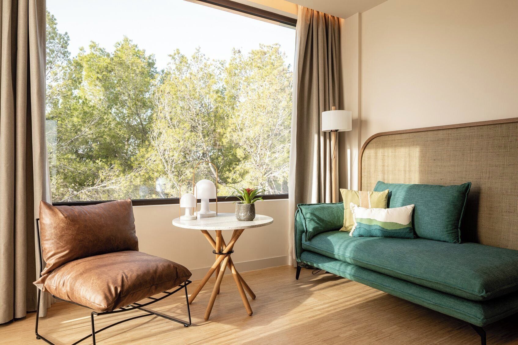 A hotel room with a leather chair, a green sofa, a small round table with a lamp, and a view of trees through the window.