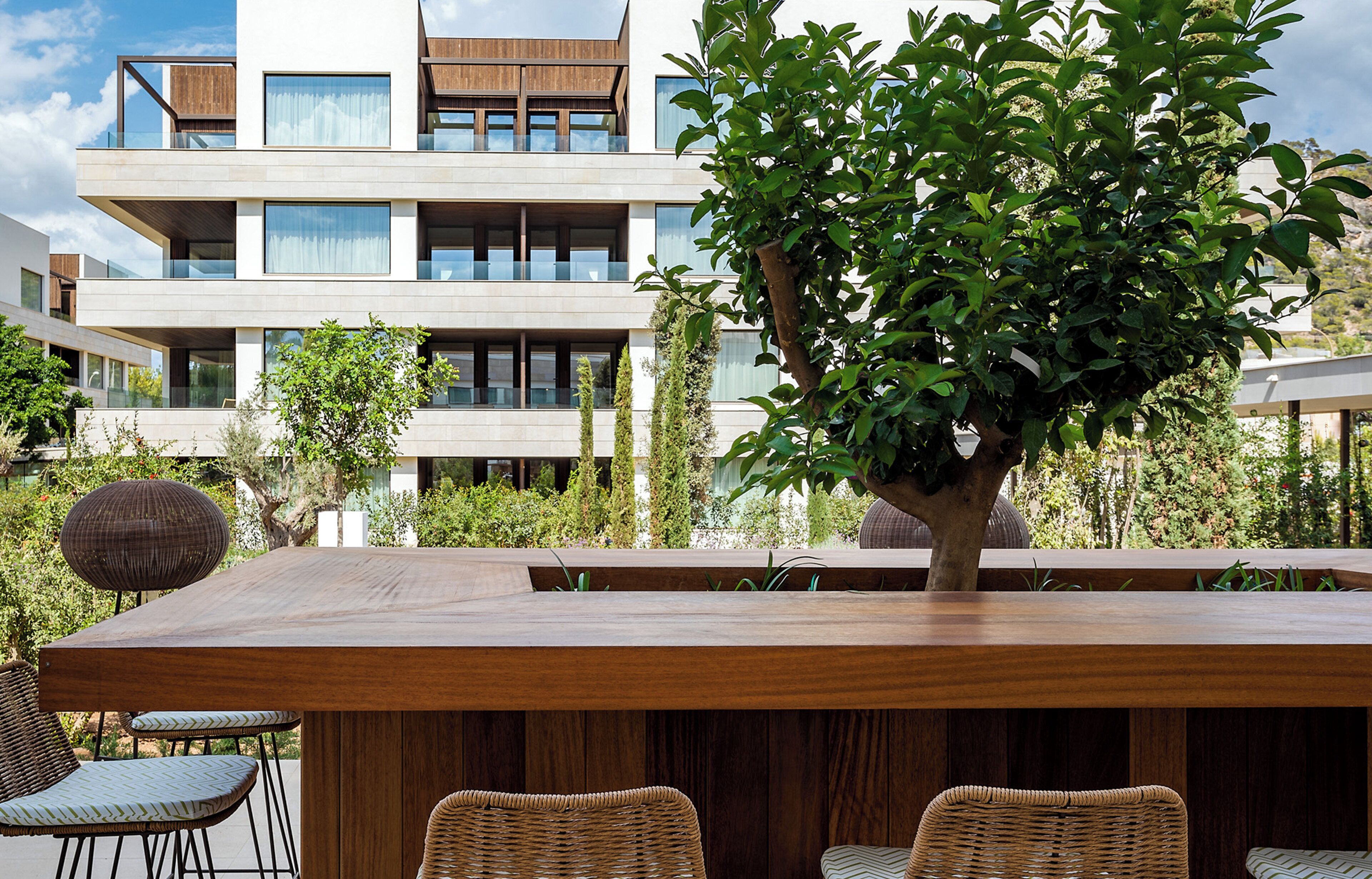 Een modern buitenmeubelgedeelte met een houten tafel en rieten stoelen, met op de achtergrond een modern gebouw met grote ramen en een boom.
