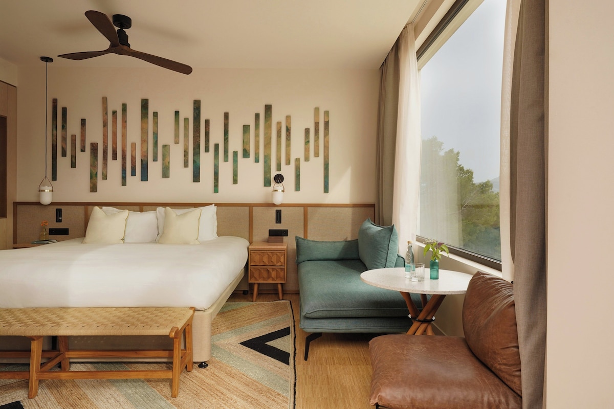 A hotel room with a bed, a sofa, a small table, a ceiling fan, and abstract wall art.