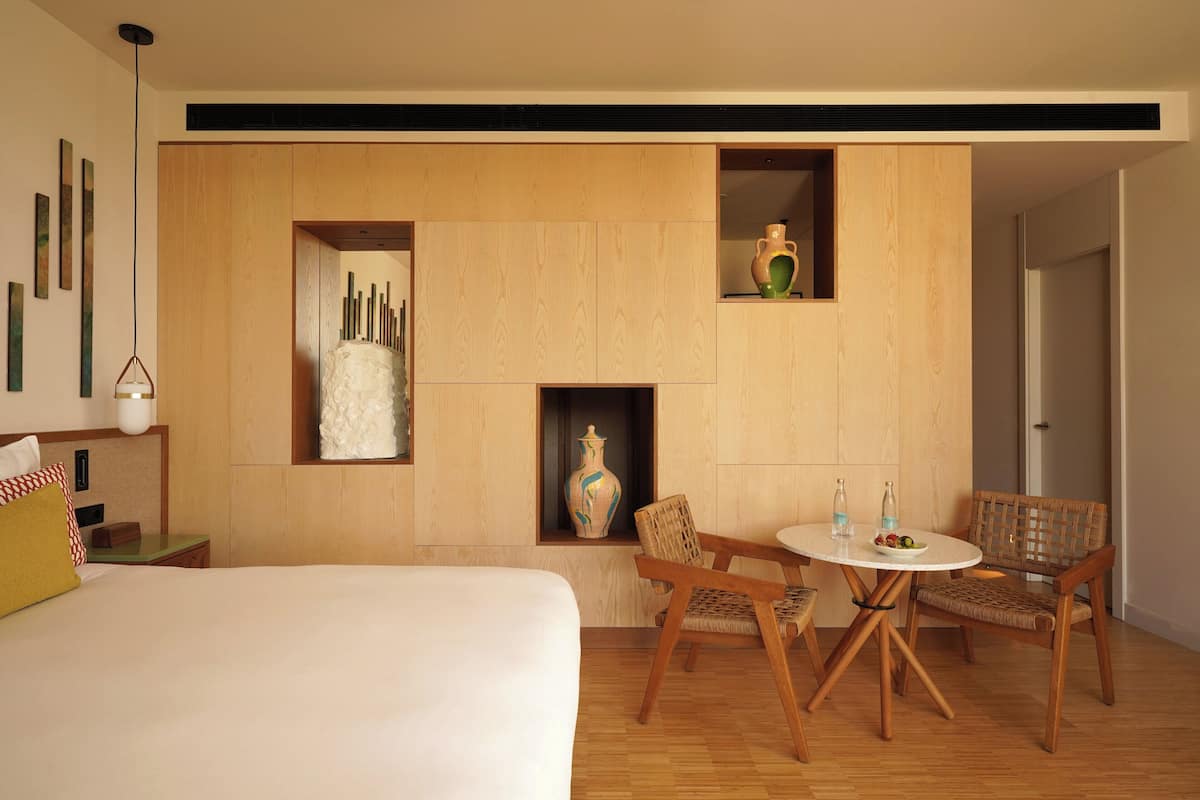 A modern hotel room with a bed, a dining area with chairs and a table, a vase, and a wall-mounted shelf with decorative items.