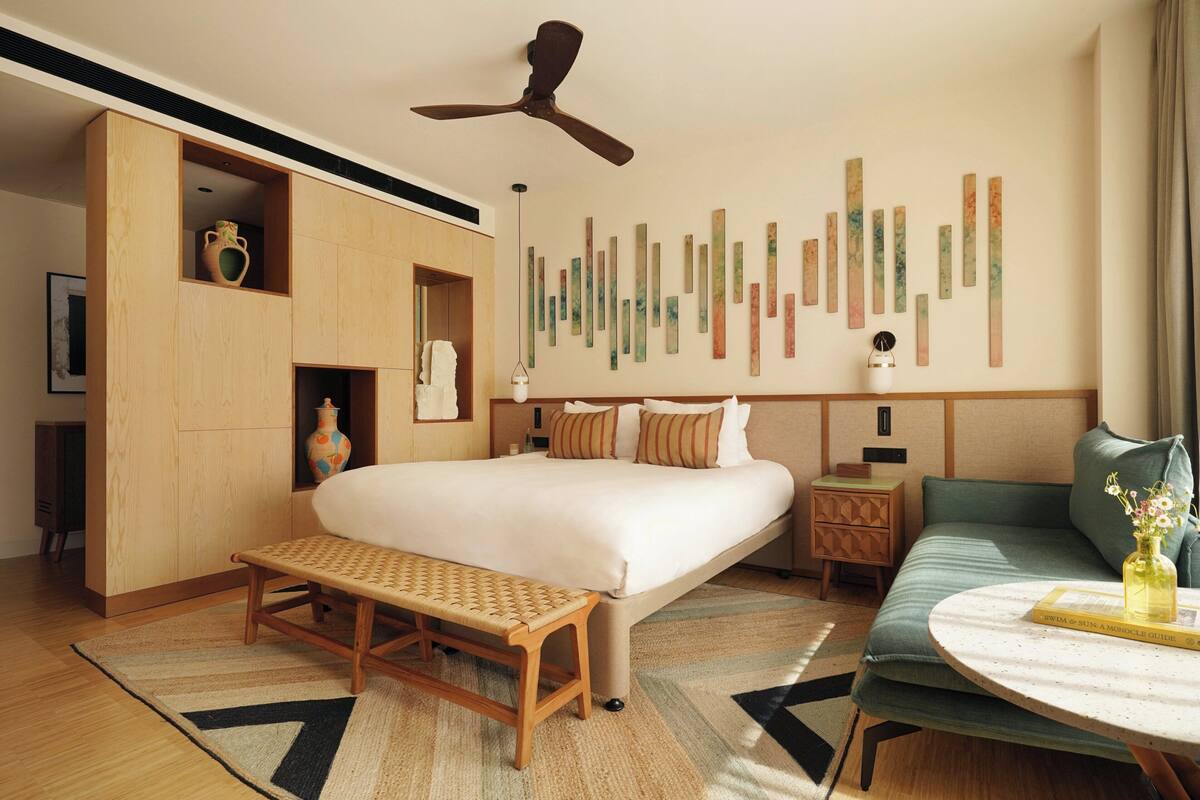 A hotel room with a large bed, a bench, a small table, a ceiling fan, and a wall with vertical wooden panels.