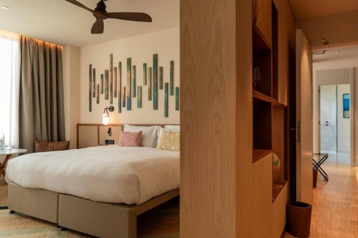A hotel room with a bed, a ceiling fan, a wall with vertical wooden slats, and a door leading to another room.